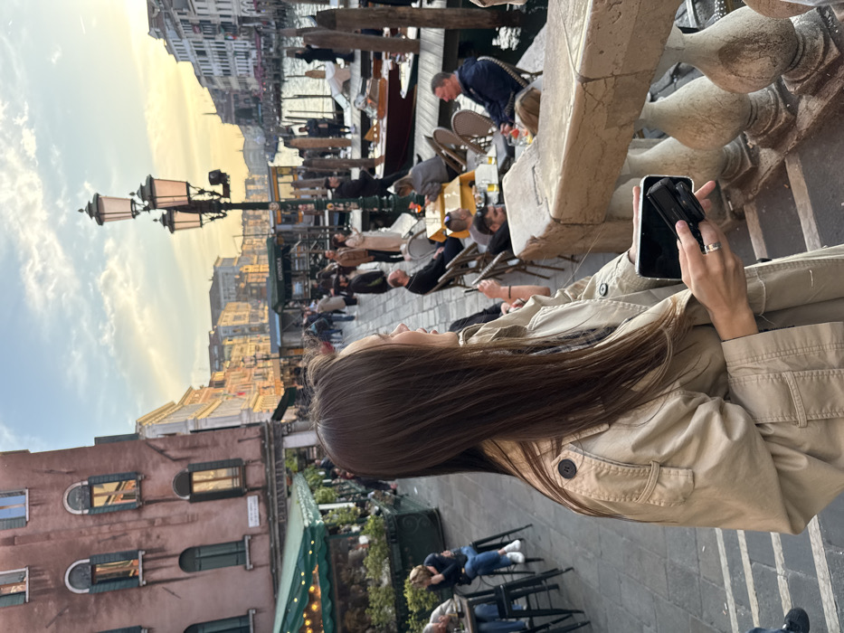 The image depicts a bustling waterside scene along the Grand Canal in Venice, Italy, during what appears to be late afternoon or early evening. In the foreground, a female subject, viewed from the upper torso, with long, straight brown hair, wears a light-colored (beige/tan) trench coat. She is turned to her right, observing the activity along the canal. Her left hand, featuring a silver ring on the ring finger, holds a smartphone horizontally, with a smaller black camera or accessory visible within its case or attached to it.
To the immediate left of the primary subject, a multi-story, reddish-brown building with multiple arched windows and dark green shutters is visible. Below this building, an outdoor dining or cafe area with green awnings is set up, where individuals are seated on stools. A light-colored stone balustrade forms the immediate foreground barrier between the viewer and the scene.
The background features a lively pedestrian promenade bordering the Grand Canal. Numerous people are present, walking or seated at outdoor cafe tables, some with yellow tablecloths, socializing and drinking. A prominent lamppost with three lanterns stands near the canal's edge. The Grand Canal itself is active, with several boats, including water taxis and gondolas, docked at wooden mooring poles. A sign displaying "GONDOLA" is visible near the waterside. Buildings of traditional Venetian architecture line both sides of the canal, receding into the distance under a partly cloudy sky illuminated by a warm, golden light.
