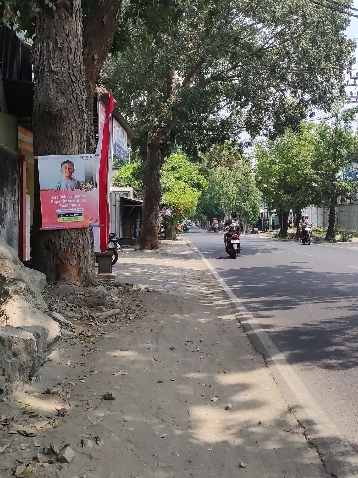 The image shows a street scene in Blitar, Indonesia. On the left side of the image, a poster is attached to a tree. The poster is for "Les Bahasa Inggris Super Interaktif" (Super Interactive English Language Classes) and features a smiling young boy. The poster also includes contact information. Adjacent to the poster, a red and white Indonesian flag is hanging.
The street itself is paved with asphalt and has a white lane marker. Several motorcycles are visible on the road. In the foreground, there is a dirt and gravel area with some rocks and debris. Trees line both sides of the street, providing shade, and the sky is bright and clear. The overall impression is of a typical urban street in a warm climate.