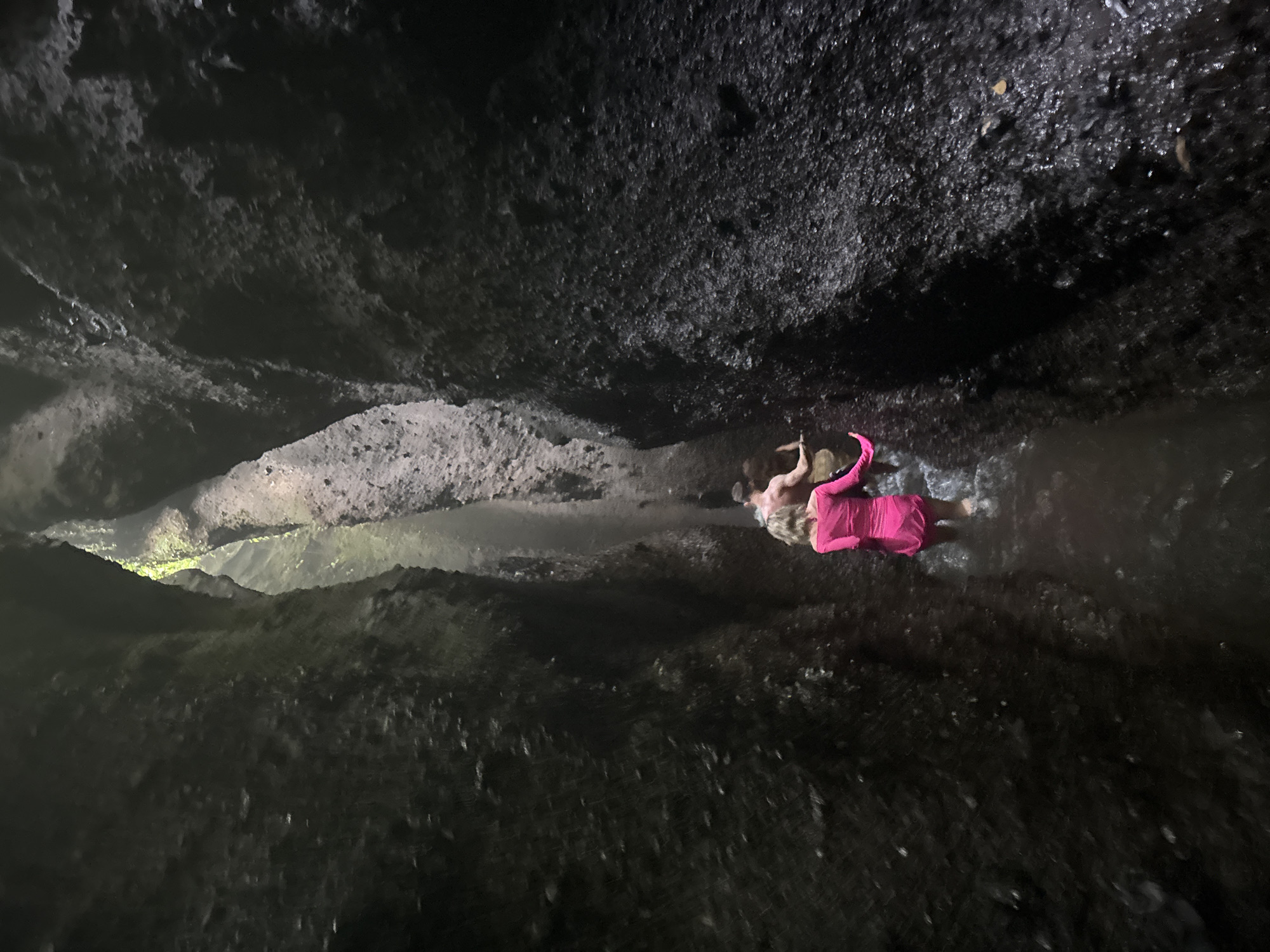 The image shows two people walking in a narrow canyon with dark, wet rock walls. The person in the foreground is wearing a bright pink dress, and the person behind them appears to be a shirtless man. Water is flowing around their legs. The canyon is dimly lit, with some light filtering in from above. This scene is likely from the Tukad Cepung Waterfall in Bangli, Indonesia, known for its unique cave-like waterfall and rock formations.