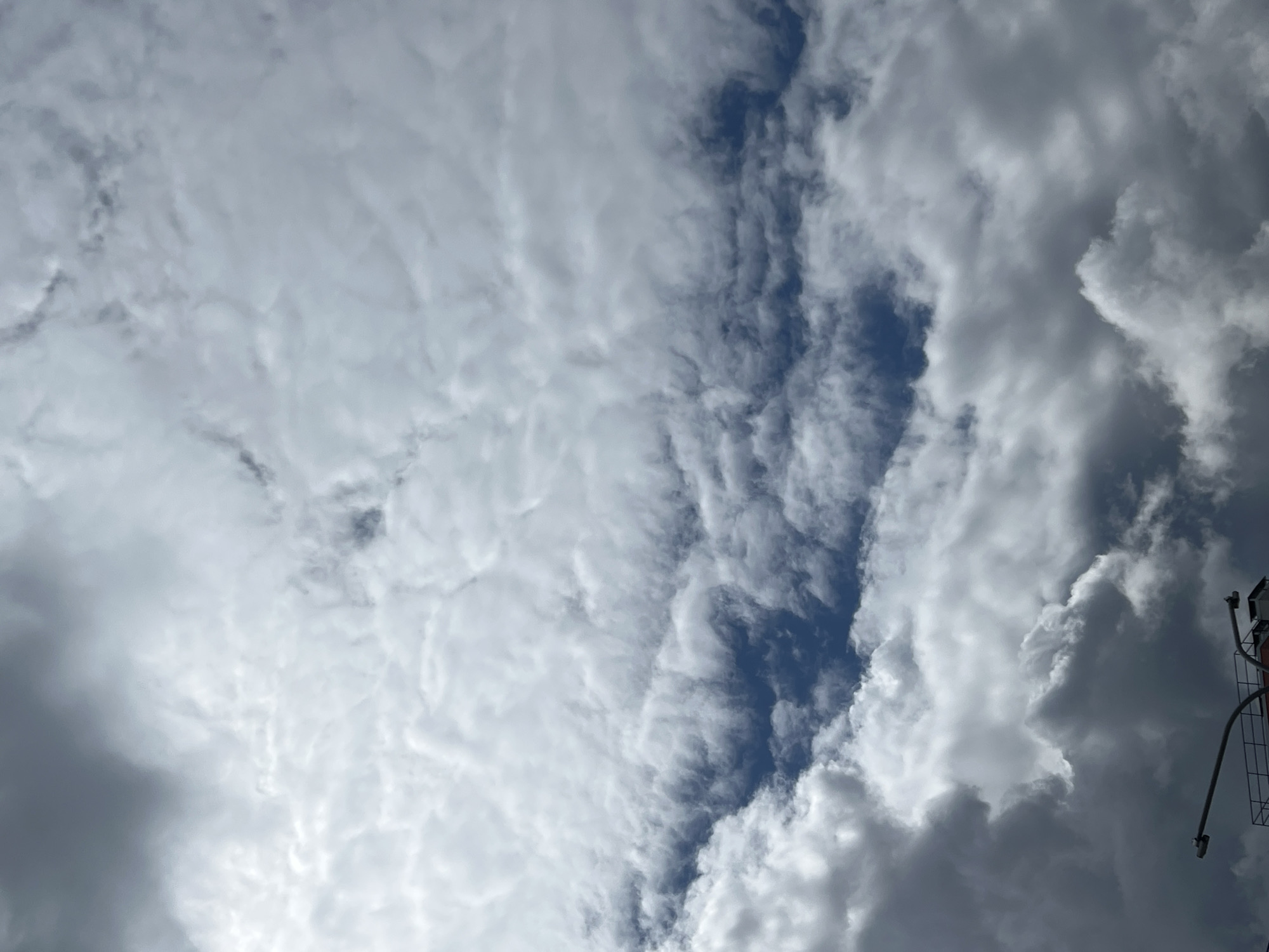The image shows a cloudy sky with a small patch of blue visible. The clouds are mostly white and gray, and they appear to be layered. In the bottom right corner, there is a partial view of a metal structure, possibly part of a building or tower, and some wires. The city of Chaikovsky, Russia, is not visibly evident in the image, but the location metadata might be related to the area the photo was taken.