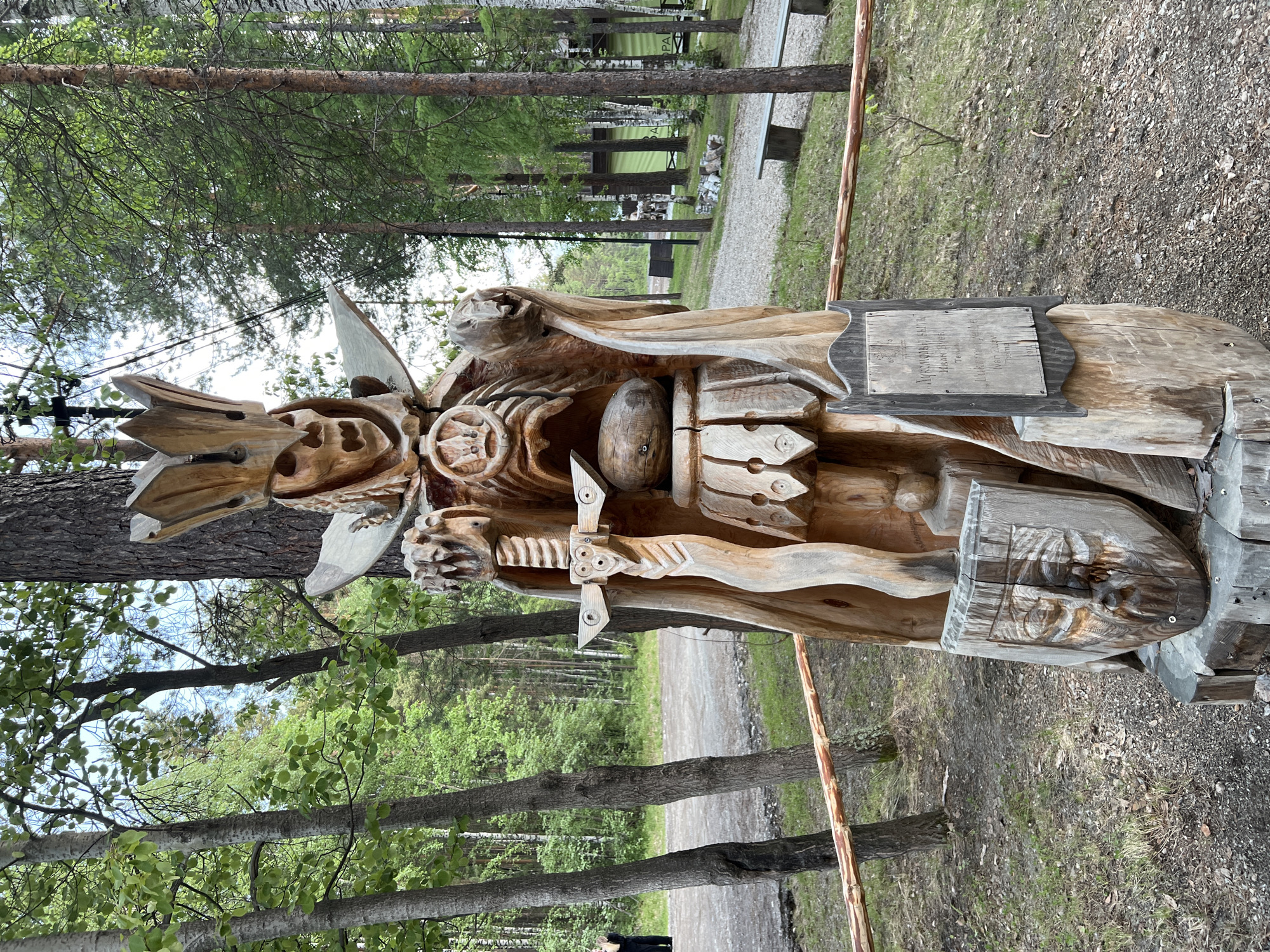 A wooden sculpture of a king or warrior figure stands in an outdoor setting, likely a park or historical site, at the Taltsy Museum, Tal'tsy, Russia. The figure is elaborately carved, featuring a crown, a skeletal face, a cape, and a sword. A shield with a carved face rests at its base. The sculpture is positioned in front of a line of trees, with a dirt path and grassy areas visible in the background. A wooden fence runs horizontally across the foreground, separating the viewer from the sculpture. The overall lighting suggests it is daytime, with overcast skies.