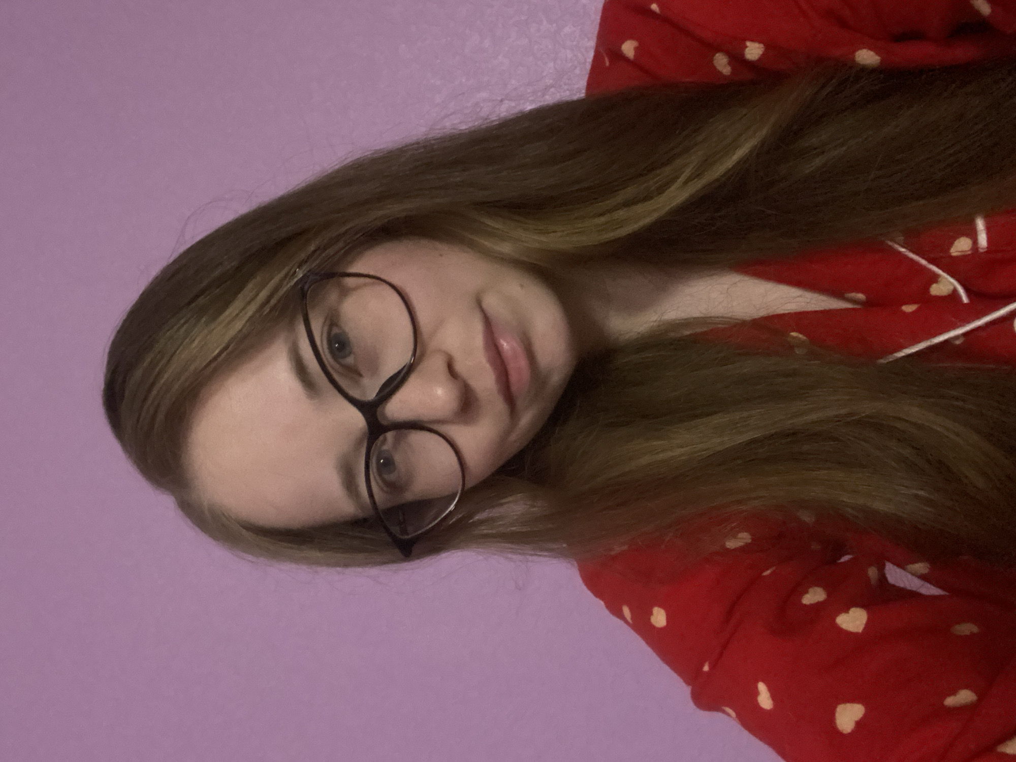 The media file displays a close-up portrait of a young woman, with her head tilted significantly to the viewer's left. She has long, straight, light brown hair with lighter highlights, which cascades around her face and over her right shoulder. She wears a pair of dark-rimmed, round glasses, behind which her light-colored eyes (appearing blue or grey) are visible, looking directly at the camera. Her lips are slightly parted in a soft, neutral expression or a subtle smile. Her skin is fair.
She is dressed in a red top, likely pajamas, characterized by small, light-colored heart patterns and thin white piping along what appears to be a collar or placket. The background is a plain, solid light purple or lavender wall, suggesting an indoor setting such as a bedroom or a casual living space.
The lighting is soft and even, indicating either indoor artificial lighting or indirect natural light, making it difficult to ascertain the exact time of day. There are no visible outdoor elements, specific location cues within the image (beyond the provided context of Saint Petersburg, Russia), or discernible weather conditions. No text is visible in the frame. The overall impression is one of casualness and relaxation.