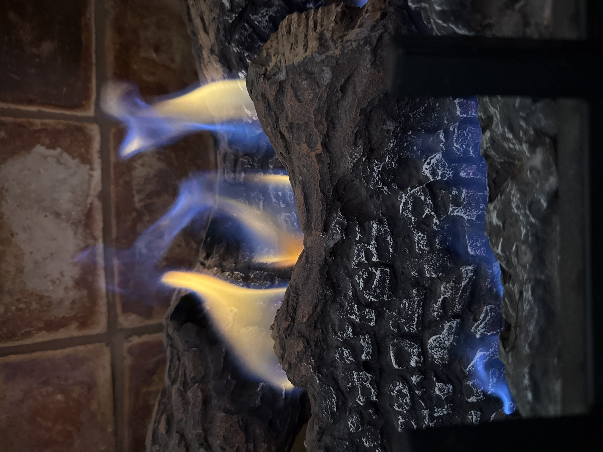 This close-up image captures an active gas fireplace, highlighting vibrant flames against artificial logs and tiled surfaces. On the left, several square terracotta-colored tiles with dark grout lines form a patterned background, suggesting a hearth or wall adjacent to the firebox. These tiles display a rustic, worn appearance.
The central and right portions of the frame are dominated by dark, textured artificial logs, likely ceramic, designed to resemble charred wood or coal. These logs feature rough, irregular surfaces with dark gray and black tones, punctuated by lighter highlights. Bright, ethereal flames, primarily blue at their base and transitioning to a vivid yellow and orange at their tips, emanate from within and around these logs. The flames appear soft and slightly blurred, indicating movement and the intensity of heat.
A dark, solid rectangular element, possibly part of a grate or the fireplace frame, is visible along the far right edge of the image. The scene is indoors, showcasing the functional operation of a gas fireplace, likely providing both warmth and ambiance. No individuals are present, and there is no visible text. While the location context of Madrid, Colombia is provided, the image itself does not contain any specific visual cues to confirm this geographical detail.