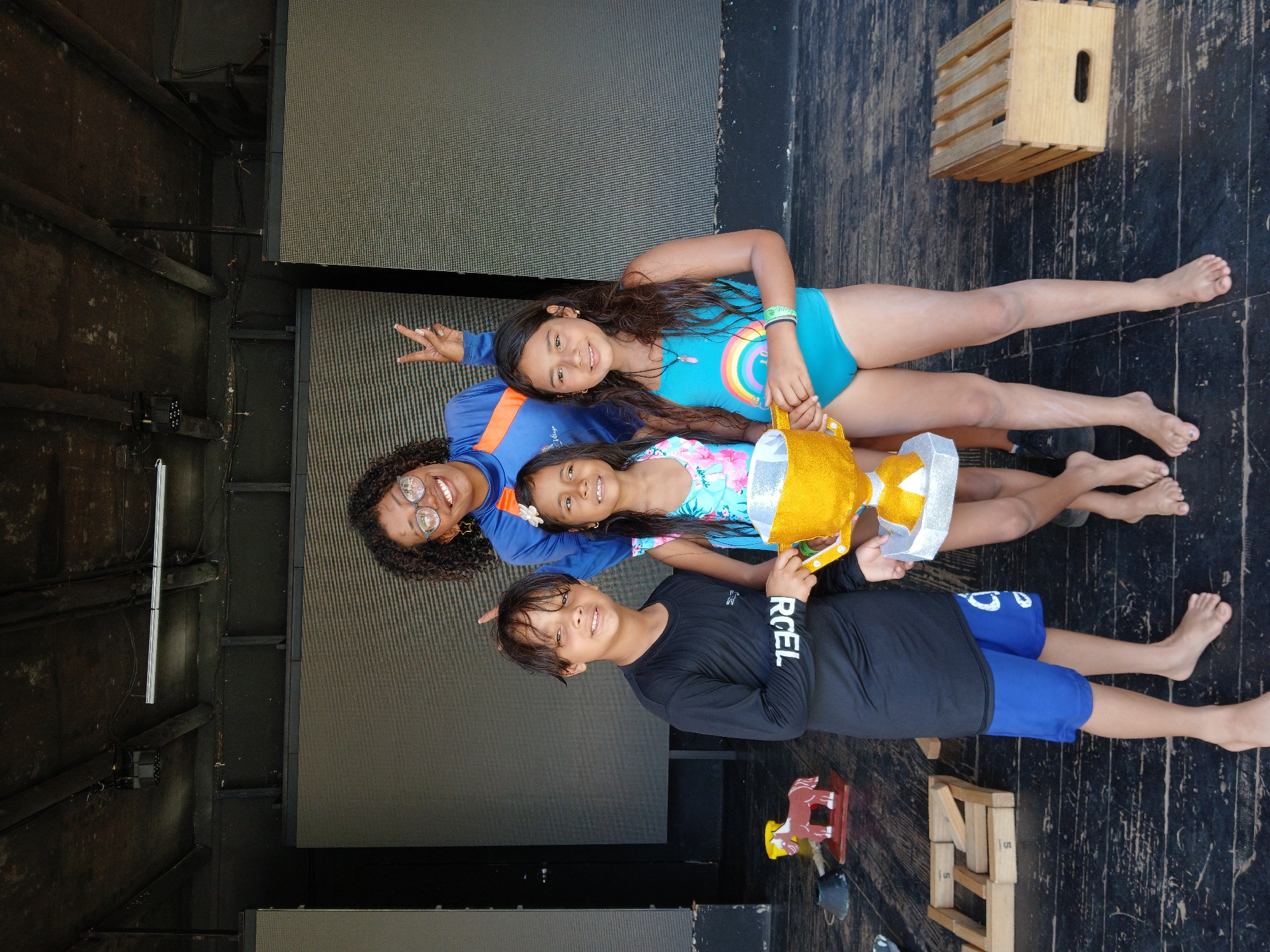 A group of four people (three children and an adult) are posing for a photo on what appears to be a wooden stage in Porlamar, Venezuela. The adult is an African American woman with glasses, wearing a blue shirt with orange stripes. The children are all barefoot. One girl is wearing a light blue bathing suit with a rainbow design, another is wearing a light blue floral print suit. The boy is wearing a black long sleeve shirt and blue shorts. The group is holding a gold and silver trophy. A wooden crate and a small rocking horse are visible in the background. The stage is set up against a large screen.