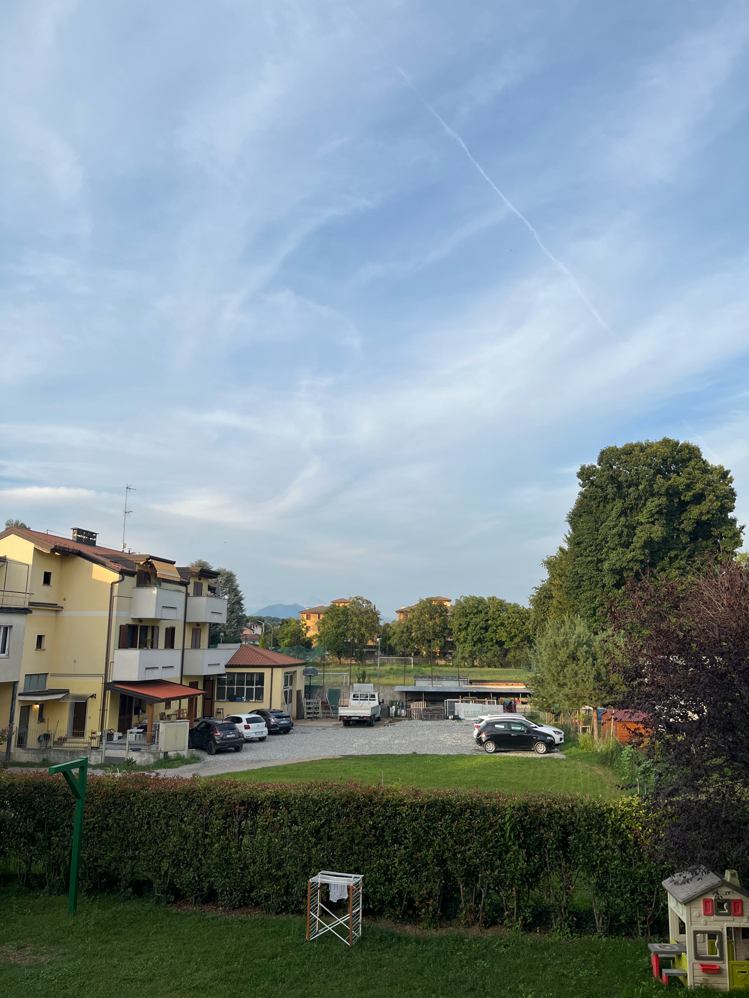 The image depicts a daytime scene in Mariano Comense, Italy. In the foreground, a well-maintained green lawn with a clothesline and a children's playhouse is visible. A dense green hedge separates the lawn from a gravel parking area. Several cars, including a white hatchback and a dark-colored SUV, are parked in this area. Behind the cars, a yellow apartment building with balconies and red-tiled roofs stands prominently. Further in the background, a large, leafy tree dominates the right side of the frame, with other trees and buildings visible in the distance. The sky above is a mix of blue with scattered white clouds, and a faint contrail stretches diagonally across it. The overall atmosphere suggests a residential area in a suburban or semi-rural setting.