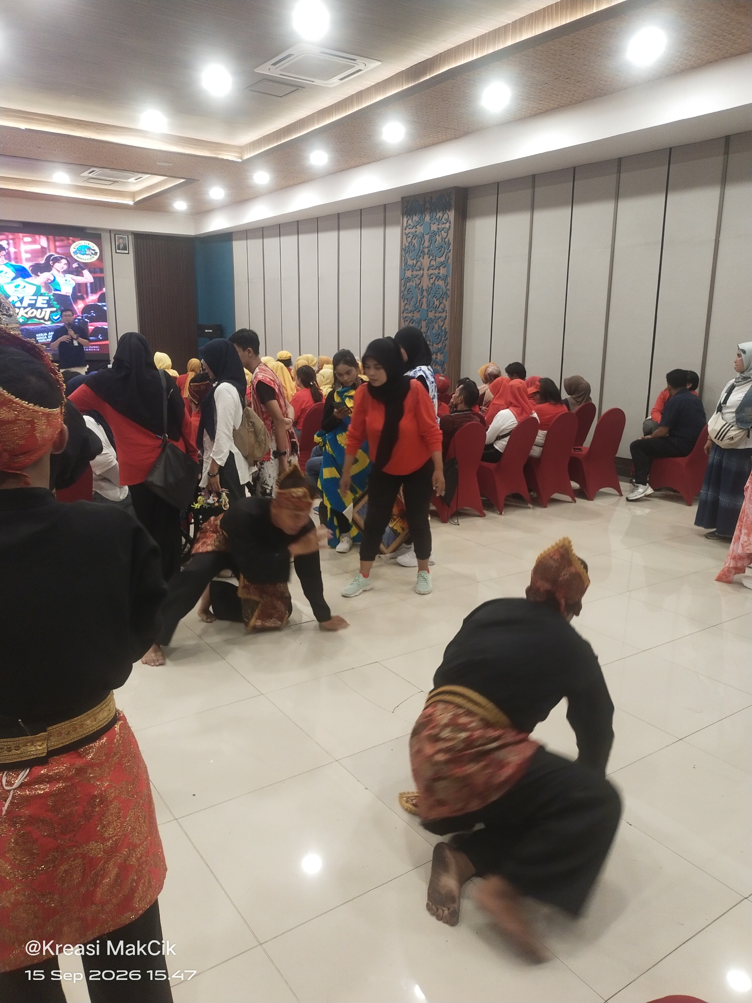 The image depicts an indoor event, likely a performance or presentation, taking place in Surabaya, Indonesia. The room is brightly lit with a tiled floor and a white, paneled wall on the right side. A large screen displaying graphics and text, including the words "LIFE" and "WORKOUT", is visible on the left. A crowd of people is seated in red chairs, observing the activity in the foreground. In the center of the floor, two individuals are engaged in what appears to be a dance or martial arts demonstration. One person is in a crouched position, wearing a black top and a red and gold patterned sarong. The other individual, also in a black top and similar sarong, is in a more dynamic pose, bent low to the ground. Several other people, dressed in a variety of traditional and casual attire, are standing or moving around the perimeter of the performance area. The overall atmosphere suggests a cultural or community gathering.