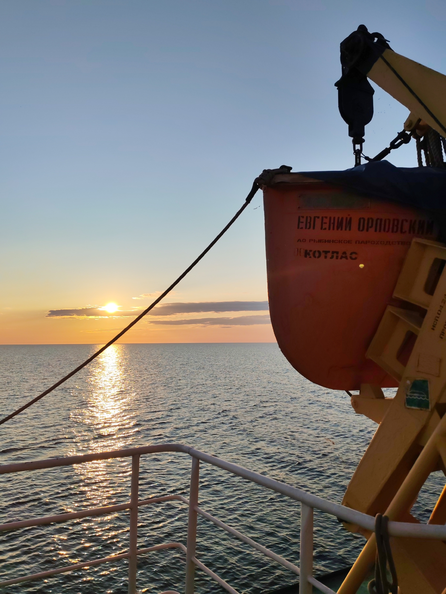 In Kakkarovo, Russia, at sunset, a ship's orange lifeboat is attached to a yellow davit. The lifeboat has Russian text indicating it is named "Yevgeny Orlovsky" and belongs to the "Rybinsk Shipping Company," with "Kotlas" also visible. The sun is low on the horizon, casting a golden path across the dark, rippling water. A metal railing in the foreground suggests the perspective is from the deck of the ship. The sky transitions from a pale blue at the top to hues of orange and yellow near the horizon.