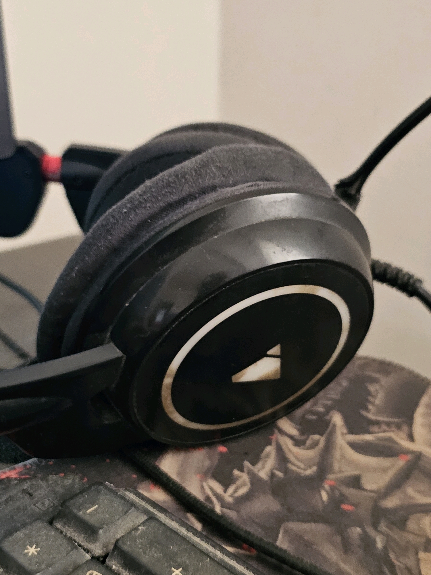 The image displays a close-up, eye-level shot of a black gaming headset. The headset is positioned on a keyboard and a patterned mousepad. The earcups of the headset are covered in black fabric, which appears slightly dusty. A white circular emblem with a triangular shape in the center is visible on the side of one earcup. The surrounding environment is out of focus, with a plain, light-colored wall in the background. The scene appears to be indoors, likely in a home or office setting. There are no people visible in the image. The specific location in the city of Batna, Algeria, cannot be determined from the visual information provided in the image.