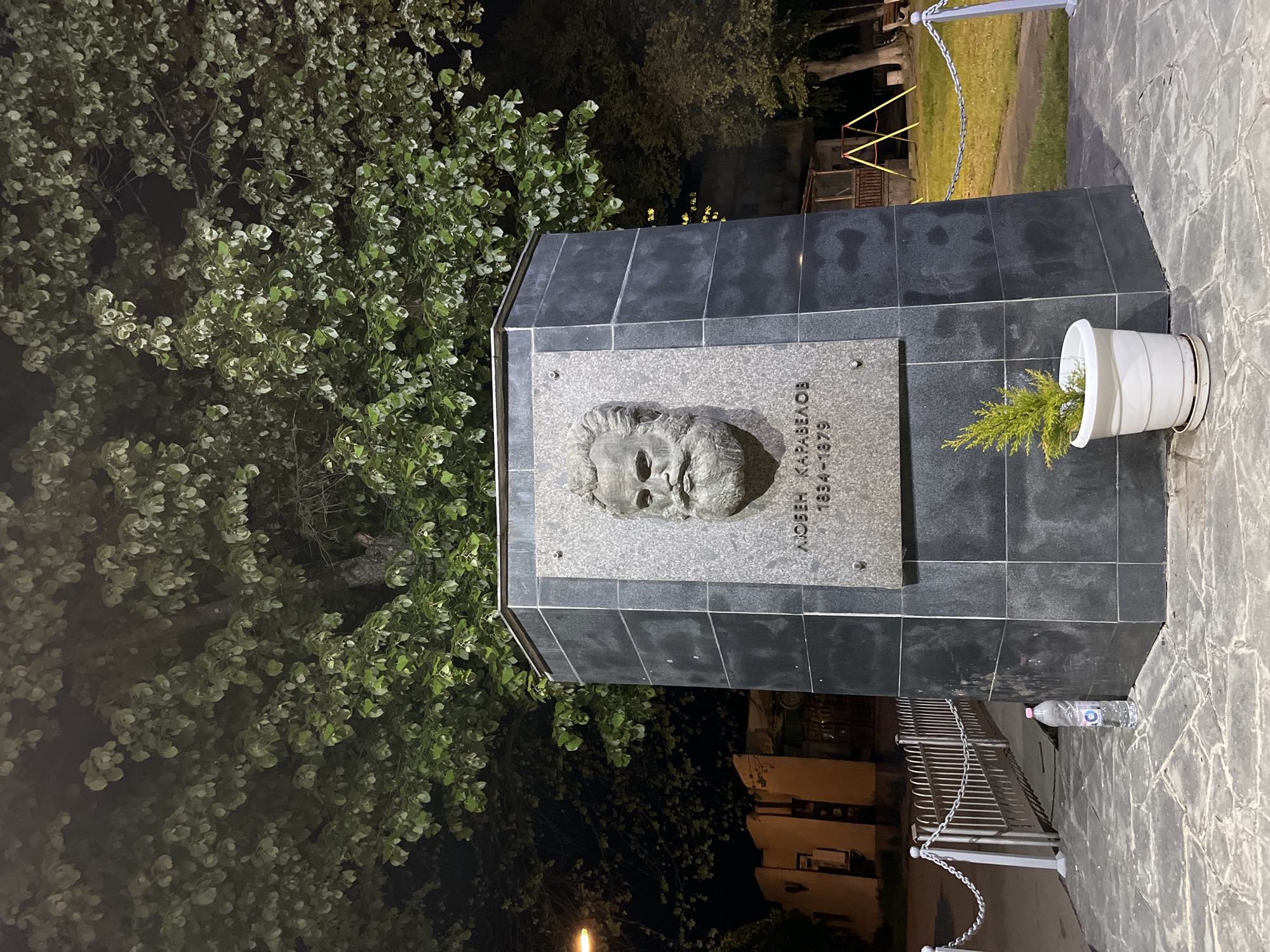 Captured at night, the image presents a monument in what can be inferred to be Karavelovo, Bulgaria, based on the inscription on the monument dedicated to Lyuben Karavelov (1834-1879). The monument is constructed from dark, possibly black, tiled stone with a lighter, stone-colored plaque affixed. The plaque bears a relief of a man's head and Cyrillic text. Next to the monument is a white pot containing a green plant. At the base, a plastic water bottle is visible. To the left, the foliage of a tree fills a portion of the frame. A chain fence and playground equipment can be seen in the background. The pavement is made of stone tiles. A building with lit windows is visible behind the fence. The lighting suggests an urban or suburban area, with illumination allowing the monument to be distinguished from its surroundings.