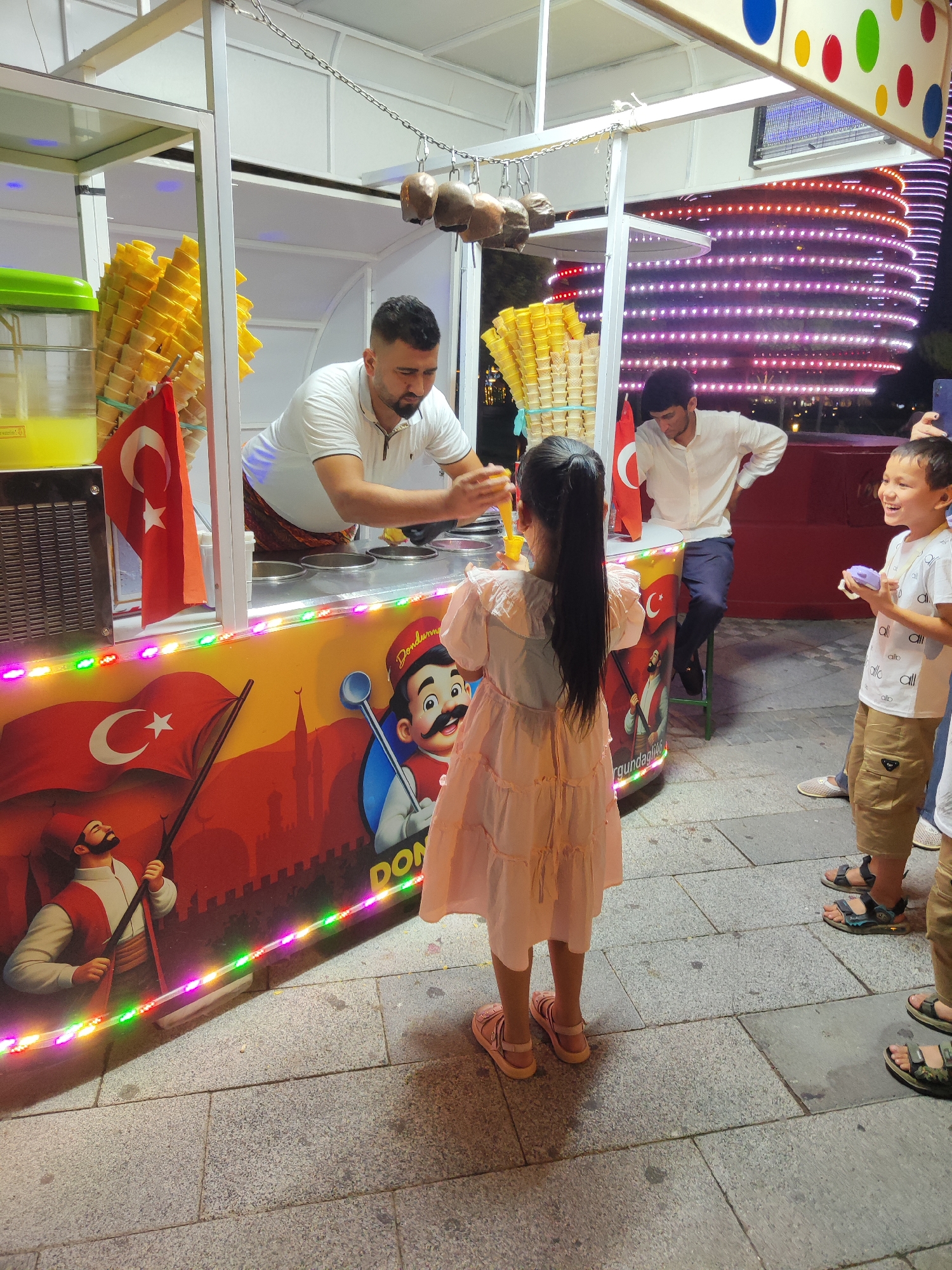 In the foreground, a young girl with long black hair, wearing a pale pink dress and pink sandals, stands with her back to the camera, facing an ice cream stand. She holds a yellow ice cream scoop. To her right, a young boy in a white t-shirt and khaki shorts, wearing blue and grey sandals, smiles and holds a small purple object. Behind him, another child's legs are visible wearing patterned sandals. The ice cream stand is decorated with colorful lights and a mural depicting Turkish flags and figures. A man in a white shirt and dark pants is serving ice cream from behind the counter. In the background, a modern building with illuminated purple and red lights is visible. The ground is paved with square stones. The scene appears to be an outdoor public space, possibly a promenade or street in Tashkent, Uzbekistan, given the presence of Turkish flags and the style of the building in the background, which resembles the characteristic architecture found in the city.
