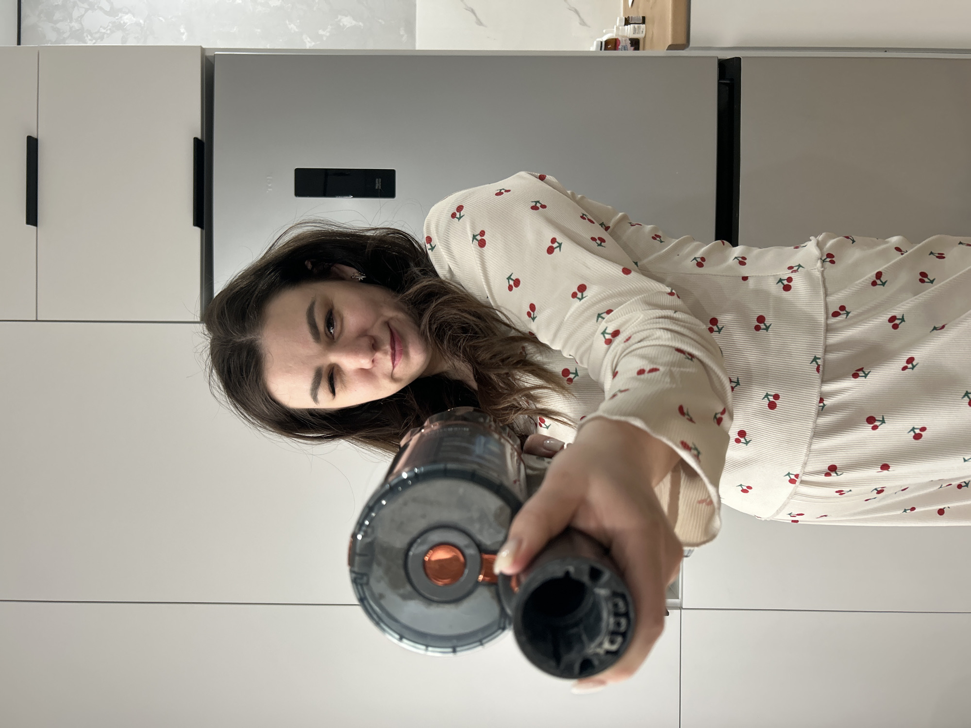 A young woman with long dark brown hair smiles and looks directly at the camera while holding a handheld vacuum cleaner. She is wearing a cream-colored, long-sleeved top adorned with a repeating pattern of red cherries with green stems. Her expression is friendly and cheerful. She holds the dark grey and black vacuum cleaner, which has a distinct orange metallic accent, pointed towards the viewer.
The background indicates an indoor setting, specifically a modern kitchen in what appears to be a home in Chaikovsky, Russia. Behind the woman, light grey or off-white flat-front kitchen cabinets with sleek black horizontal handles are visible. To her right, a built-in stainless steel or grey-colored refrigerator is integrated into the cabinetry. The refrigerator features a rectangular black control panel or display, and the faint "TCL" brand logo is visible on its surface. Above the cabinetry, a light-colored, possibly marble-patterned backsplash or wall is present, with some small bottles or jars subtly visible on a shelf in the top right corner. The overall aesthetic of the kitchen is clean and contemporary.