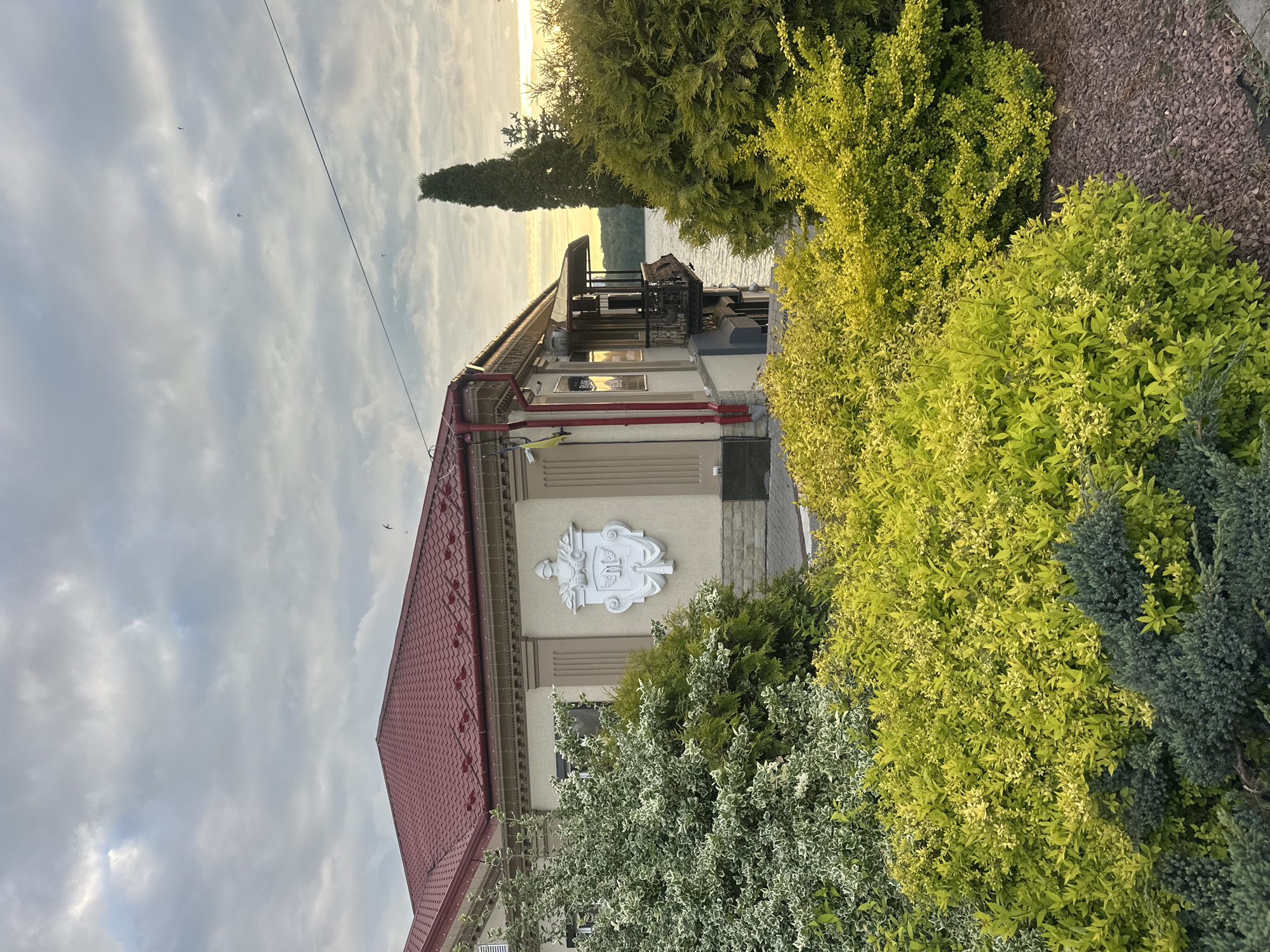 The image depicts a building with a red tile roof and a cream-colored facade, surrounded by lush greenery. A white emblem is mounted on the building's wall. A balcony with iron railings is visible, offering a view of a lake and a tree line in the distance under a cloudy sky. The location is Ternopil, Ukraine, as indicated by the emblem. There are birds in the sky. The vegetation includes green and yellow shrubs.
