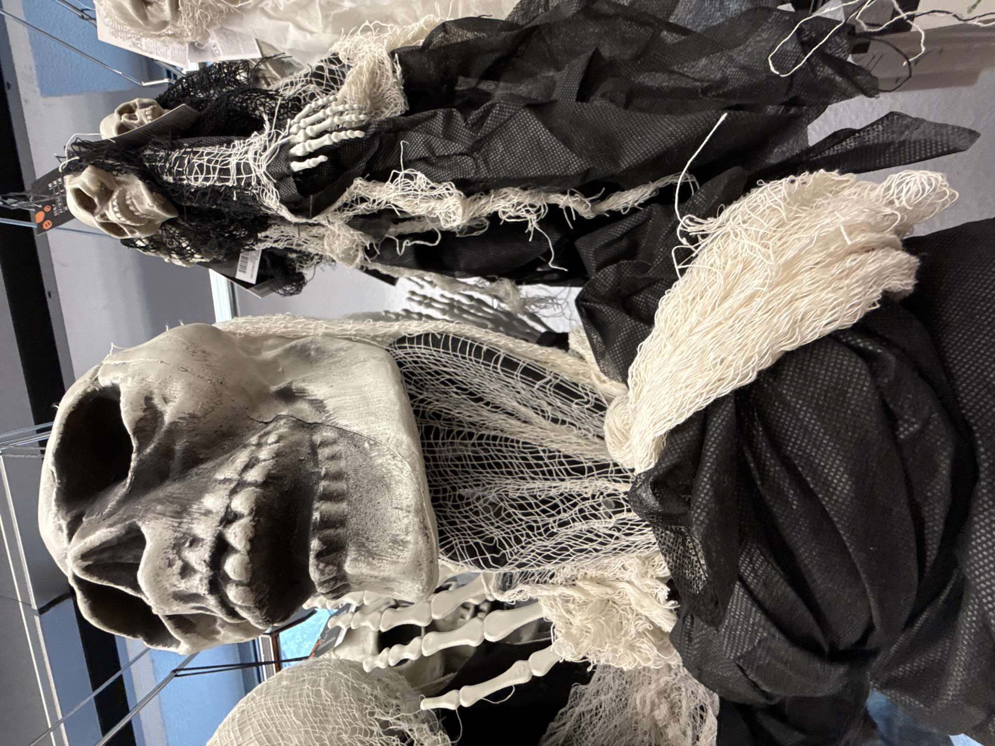 The image shows a close-up of several Halloween decorations, specifically skeletal figures. The primary focus is on a large, white skull in the foreground, with a gaping mouth and visible teeth. This skull is draped with tattered white netting and black fabric. Behind it, to the right, are two smaller skeletal figures, also adorned with black netting and white stringy material, with visible plastic hands. To the far left, another skeletal figure wrapped in white netting is partially visible. The background is blurred but suggests an indoor setting with overhead lighting and possibly shelving or displays. There is no indication of a specific location in Bad Lauterberg im Harz, Germany.