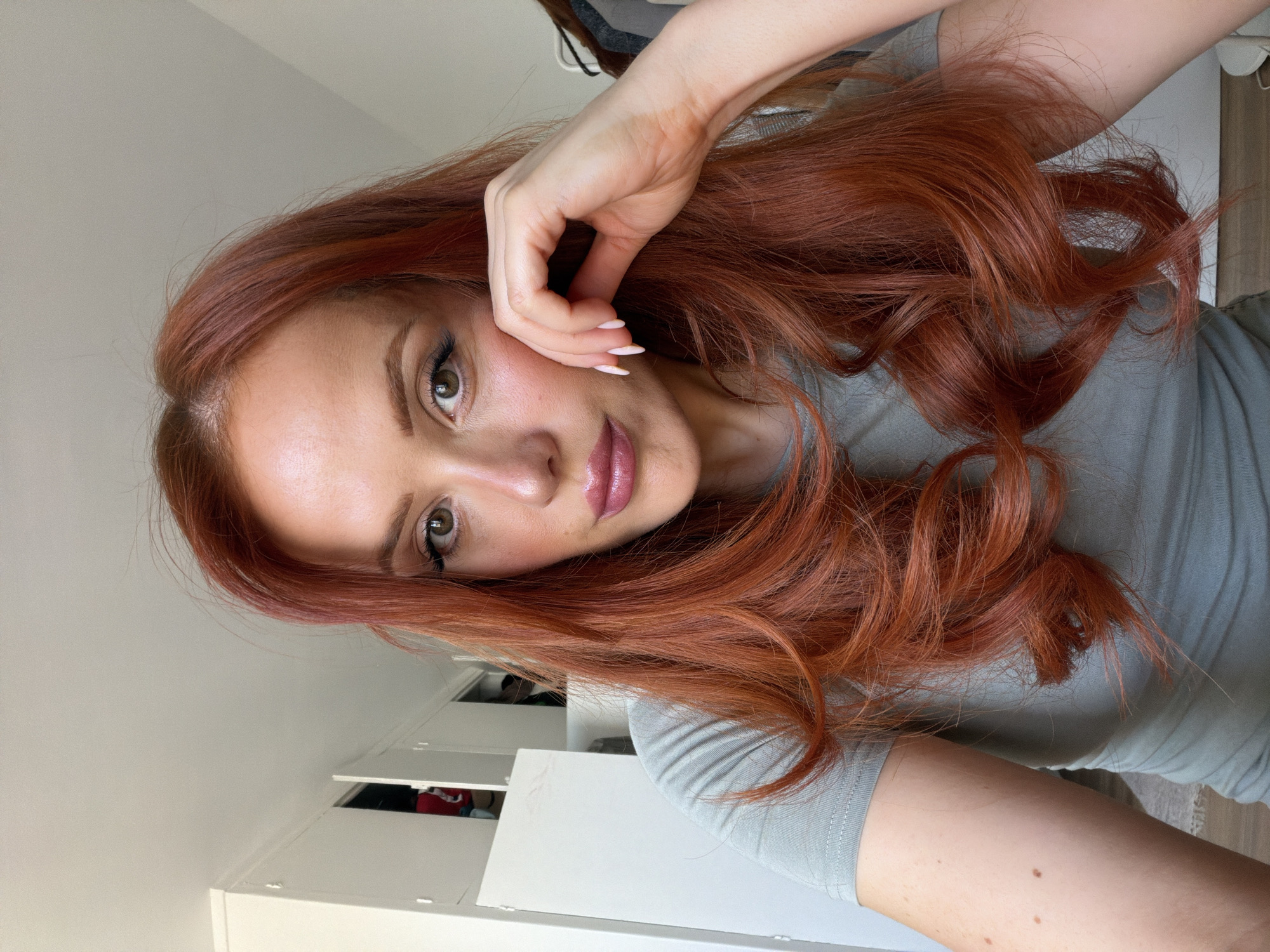 A woman with long, wavy, auburn hair is captured in a medium close-up shot. She is wearing a light grey t-shirt. Her right hand is resting on her cheek, with her fingers gently touching her jawline. She has light makeup on, including eyeliner and lipstick. The background appears to be indoors, with white cabinets visible on the left and part of a grey garment on the right. The lighting suggests a well-lit room, possibly during the daytime. There is no indication of the location being Paris, France.