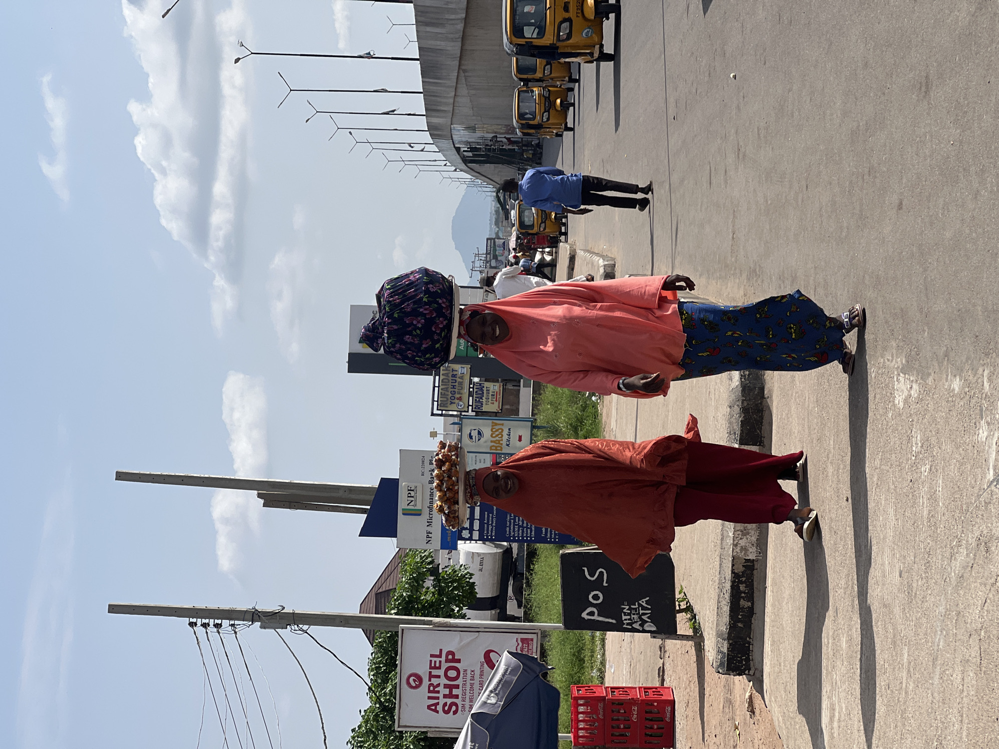 The image depicts two women walking on a street in Jalingo, Nigeria, carrying goods on their heads. The woman in the foreground wears an orange headscarf and a matching orange and red outfit, with a basket of food items balanced on her head. Behind her, another woman in a pink hijab and blue patterned skirt also carries a large, dark-colored bundle on her head.
The scene appears to be a busy street with several yellow tricycles, commonly known as "Keke Napep," lined up on the road. A concrete overpass stretches across the background, with streetlights visible along its edge. Signs for various businesses are present, including "Airtel Shop," "NPF Microfinance Bank Plc," and food-related establishments like "RUFAIDAH YOGHURT & FURA" and "BASSY Kitchen." A "POS MTN=ATEL DATA" sign is also visible, suggesting a point-of-sale service for mobile data.
The weather seems fair with a bright, sunny sky and scattered clouds. The sunlight casts long shadows on the paved road, indicating it is likely daytime. The women's expressions are neutral or slightly smiling, suggesting a normal day of commerce. The overall impression is a glimpse into daily life and street vending in a Nigerian city.