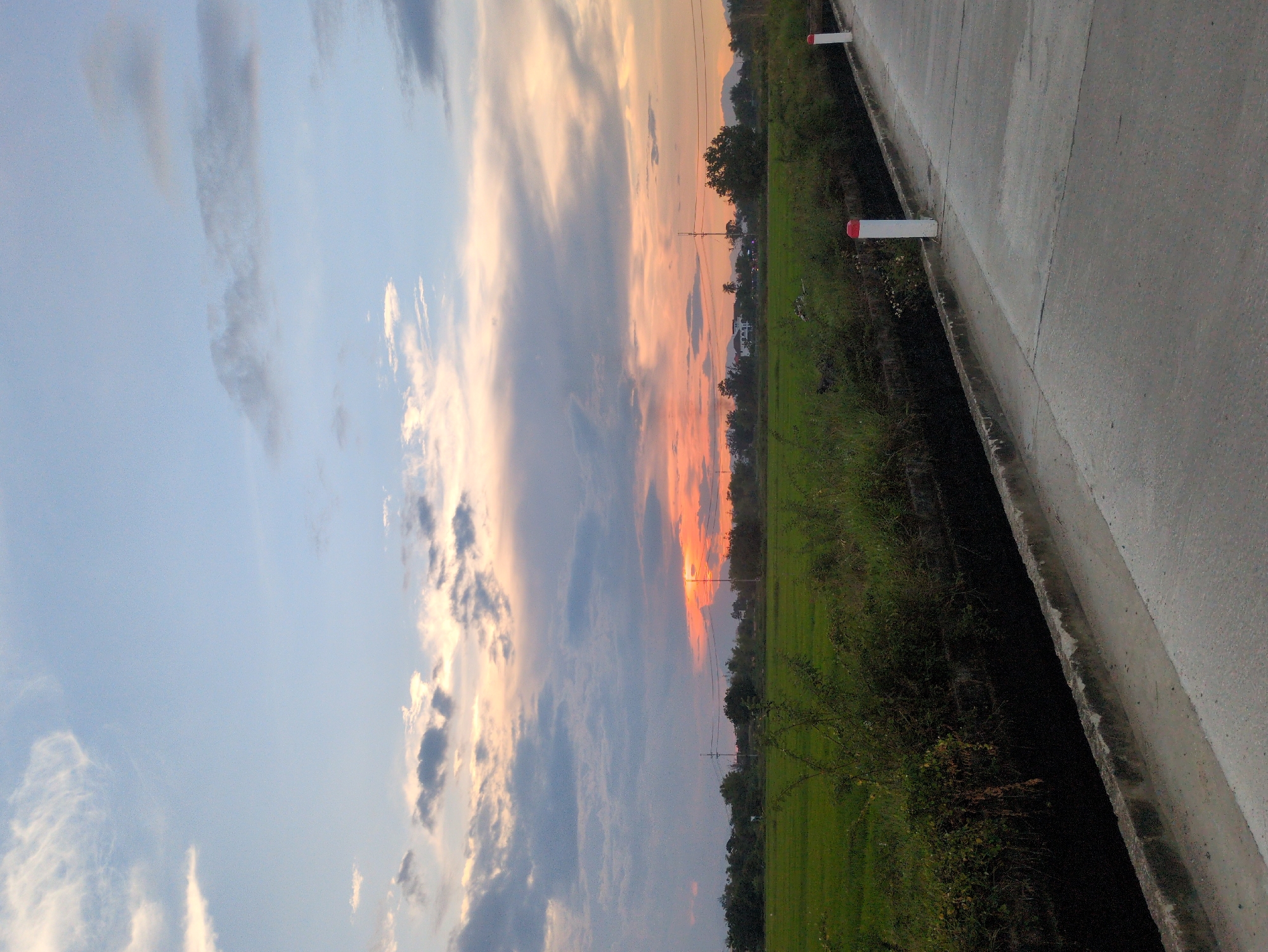 The image depicts a rural landscape in Hoi An, Vietnam, at dusk. A concrete road occupies the foreground, curving towards the right of the frame. Along the left side of the road is a narrow, dark drainage ditch bordered by a concrete edge. Beyond the ditch, a lush green rice field stretches into the distance, dotted with trees and vegetation. In the background, a line of trees separates the fields from more distant structures and mountains. The sky above is a blend of soft blues, grays, and oranges, with scattered clouds illuminated by the setting sun, casting a warm glow over the scene. Two white posts with red tops are visible along the edge of the road, marking its boundary. There are no people visible in the image. The overall atmosphere is tranquil and serene, capturing the beauty of the Vietnamese countryside at twilight.