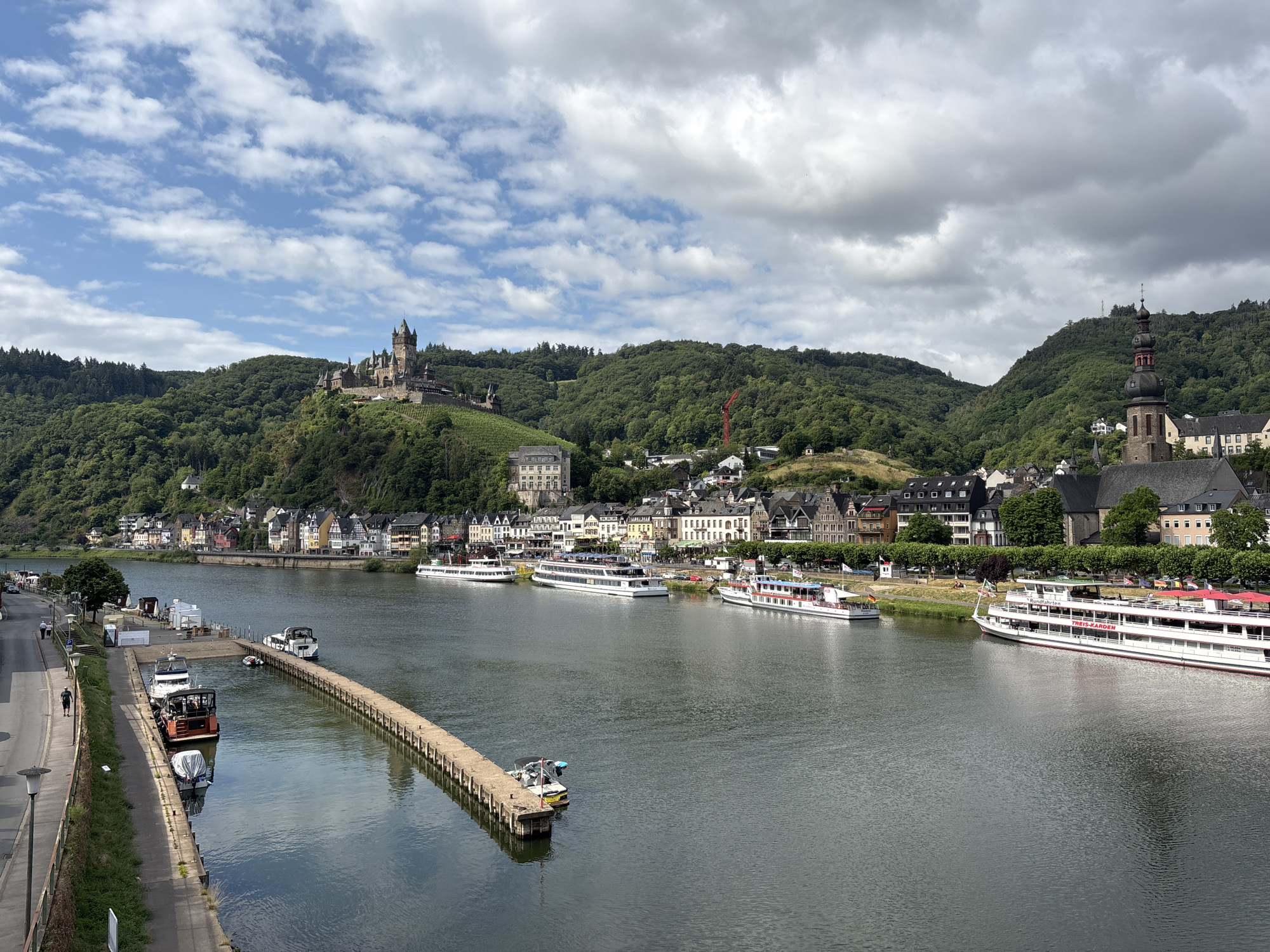 This is a high-angle, wide-shot, eye-level photograph of the Moselle River flowing through Cochem, Germany. On the left bank, a paved walkway with streetlights runs parallel to the river, and several people are walking. Adjacent to the walkway is a pier with a long, concrete jetty extending into the water, where multiple boats are docked, including a small, red and orange boat with a cover. Across the river, a picturesque town with colorful buildings lines the waterfront, backed by lush green, tree-covered hills. Dominating the left side of the frame is the Reichsburg Castle, perched atop a hill. On the right side of the town, a church spire with a dark, onion dome rises above the rooftops. Several large passenger boats are moored along the river, with one prominently displaying "Rundfahrten" and "TREIS-KARDEN" on its side. The sky is a mix of blue with scattered white clouds.