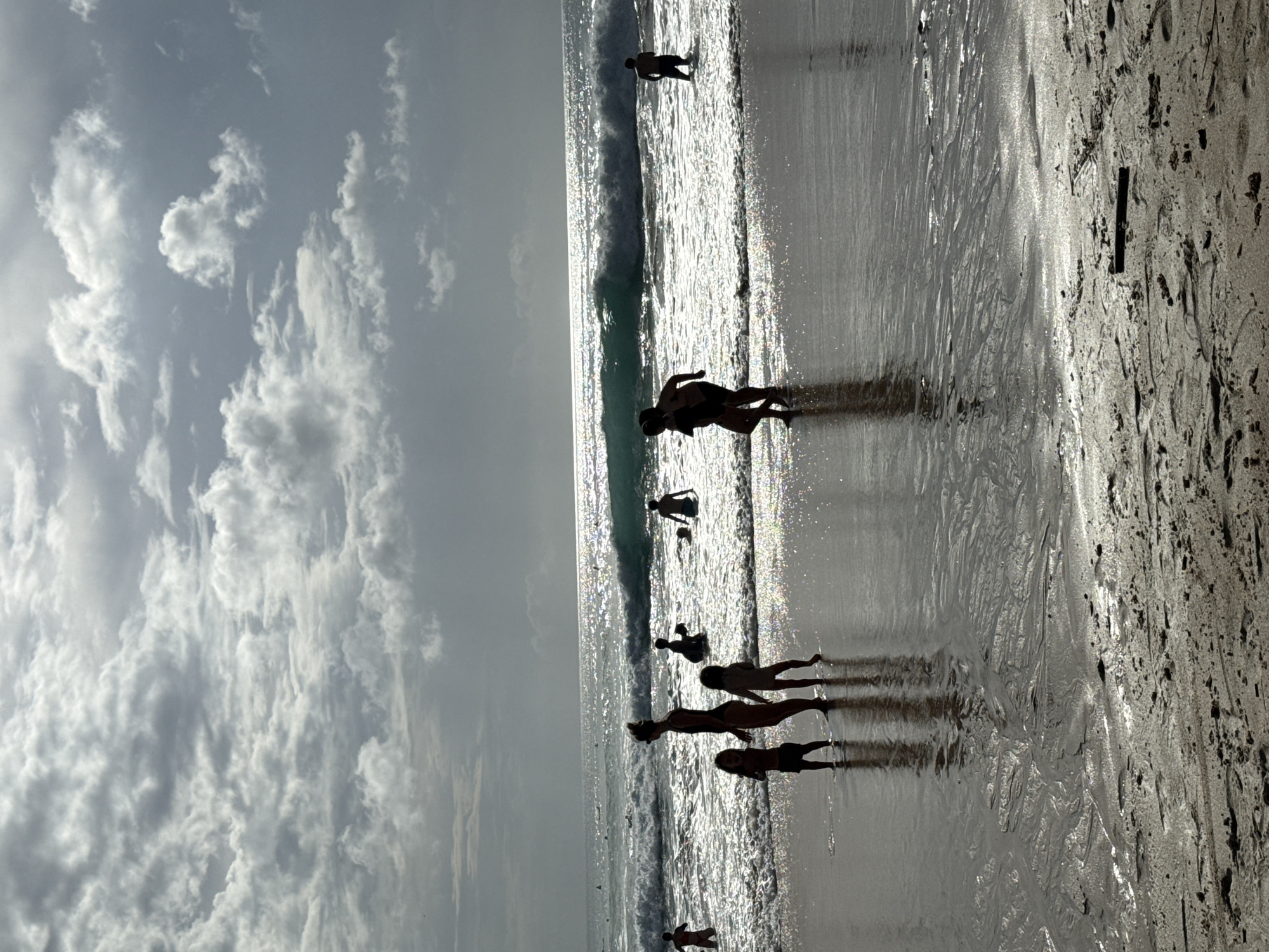The image depicts a beach scene, likely at sunrise or sunset, characterized by strong backlighting that creates dramatic silhouettes. The upper portion of the frame is dominated by a cloudy sky, with patches of bright white cumulus clouds set against a softer, hazy grey background.
The horizon line is visible, separating the sky from the ocean. The water surface intensely reflects the sun, appearing as a shimmering expanse of bright silver and white, with visible breaking waves adding texture.
Several individuals are captured in silhouette against this luminous backdrop. In the foreground, a group of four people – appearing to be two adults and two children – stand at the water's edge on the wet sand. Their long shadows stretch across the reflective surface. Further out in the water, more indistinct silhouetted figures are visible, seemingly wading or playing in the waves. The wet sand in the immediate foreground shows ripples and darker, irregular patterns.
The overall mood is one of leisure and tranquility. The strong contrast between the dark silhouettes and the sparkling water is a notable aesthetic detail. No text is visible within the image. The location context provided is Kuta Selatan, Indonesia.