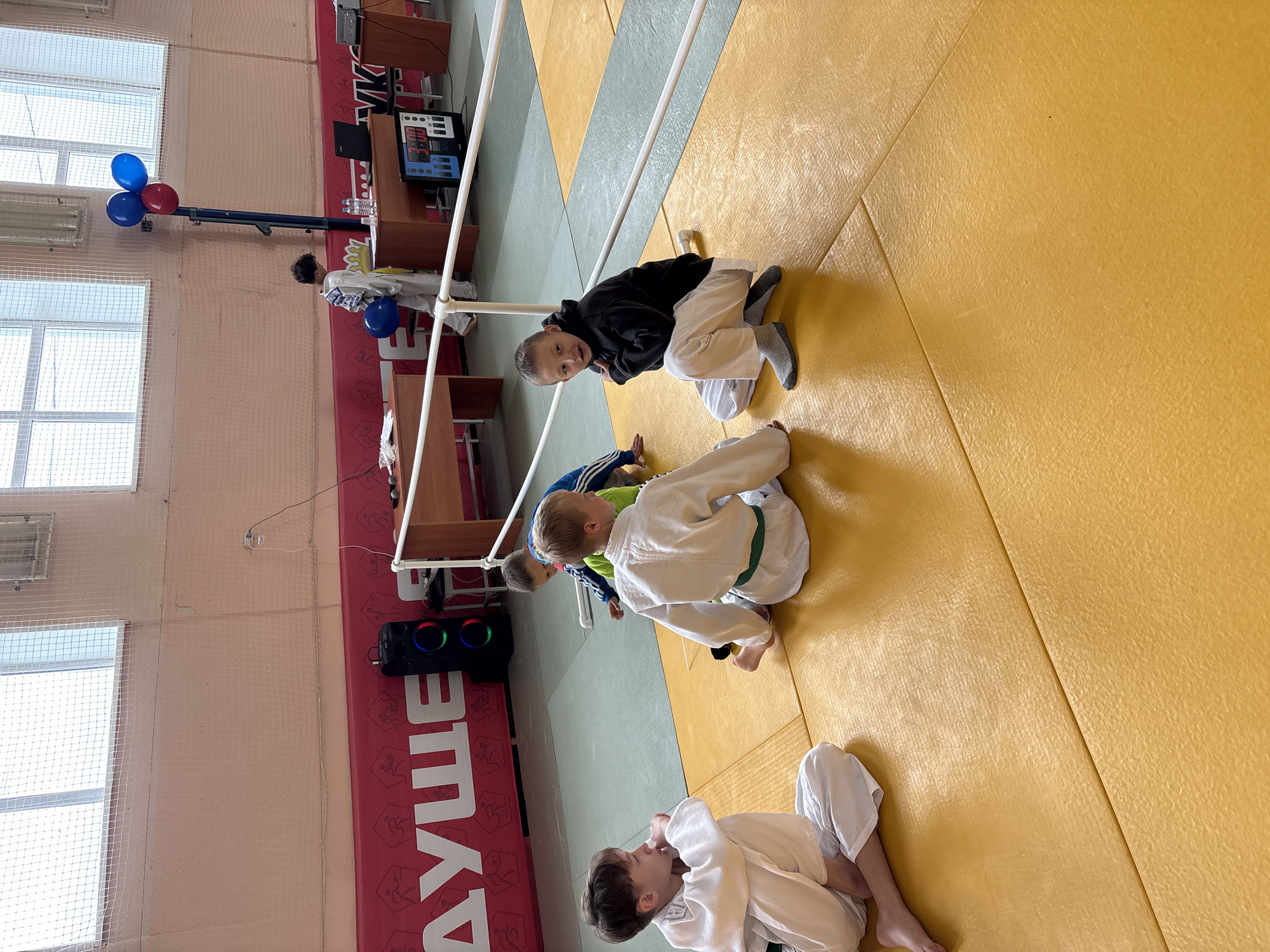 This image captures an indoor sports facility, likely a dojo or gymnasium, in Kuyeda, Russia, showing a break or administrative area during a youth sports event. The foreground features a yellow tatami mat floor with a section of grey-green matting visible further back, separated by a white boundary line and a low white railing.
Five children are present in the scene. In the left foreground, a boy with light brown hair, wearing a white judogi with a green belt, sits cross-legged, facing right, with his left hand near his chin. Adjacent to him, a second boy, also in a white judogi with a green belt, sits on the mat, facing left with legs extended. To the right, a third boy with dark hair, wearing a black hooded sweatshirt and white pants with grey socks, is in a low crouch, looking towards the camera with an open mouth. Partially obscured behind the second boy, a fourth boy in a blue and green tracksuit is kneeling on the mat, facing away. In the background, near the administrative tables, a fifth boy with dark curly hair, wearing a white judogi, stands holding a blue balloon.
The background wall is painted a light peach color. A red banner with white lettering and stylized athletic figures runs along the lower portion of the wall. Partially visible text on the banner includes "ДУШЕ" and "УК". High on the wall are large, multi-pane windows covered by a fine white mesh or netting.
An administrative area is set up with two light-colored wooden tables. On the right table, a black laptop and water bottles are visible. A digital scoreboard, predominantly black with blue and white elements and displaying "W" and "W", rests on the floor near this table. Between the tables, a blue vertical pole is topped with three balloons: two blue and one red. On the left table, small dark objects resembling microphones are present, and a tall black speaker with prominent circular blue, green, and red LED lights is positioned on the floor nearby. The children on the mat appear to be resting or observing, while the boy in the black hoodie looks directly towards the viewer.