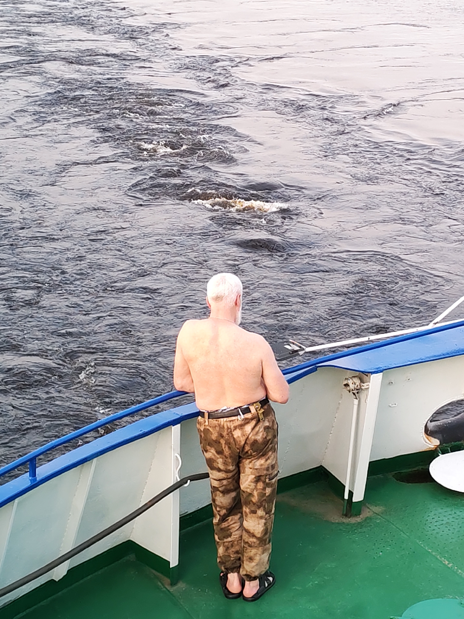 A shirtless man with white hair and a gold necklace stands on a boat, facing away from the camera, and looking out over a body of water. He is wearing patterned pants, a black belt, and dark sandals. The boat has a blue and white railing. The floor of the boat is green. The water is dark and choppy. The city is Unknown, Unknown.