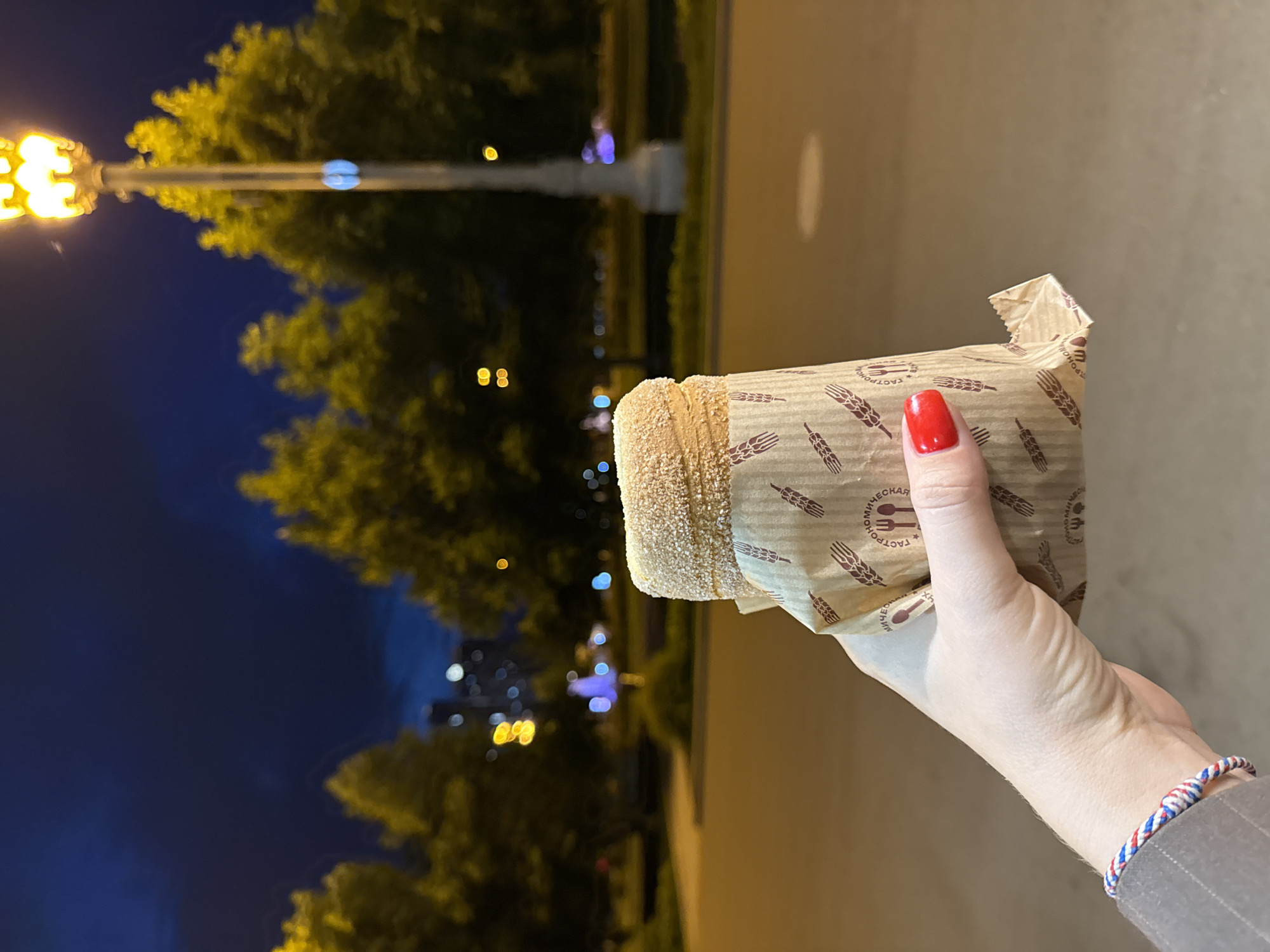 The image shows a close-up of a person's hand holding a cylindrical, sugar-coated pastry, resembling a Trdelník or similar spit cake, at night. The setting appears to be an urban or park-like area in Moscow, Russia.
The pastry, golden-brown and covered in a sparkling layer of sugar crystals, is partially wrapped in brown paper. The wrapper is adorned with a repeating pattern of stylized wheat stalks and a logo featuring a fork and knife. Visible Cyrillic text on the wrapper clearly reads "ГАСТРОНОМИЧЕСКАЯ" (Gastronomic), providing a strong location cue to Russia.
The hand, likely female, displays vibrant red nail polish on the fingernails. A braided bracelet, featuring red, white, and blue colors, is worn on the wrist, peeking out from under a grey, possibly striped, sleeve.
The background is dark, indicating nighttime. Bright yellow streetlights cast a glow on lush green trees, creating a warm contrast with the deep blue sky. Blurred bokeh lights in various colors (yellow, blue, white) are visible in the distance, suggesting other urban elements like buildings, vehicles, or streetlights. The scene depicts a casual moment, perhaps enjoying a treat during an evening walk.
