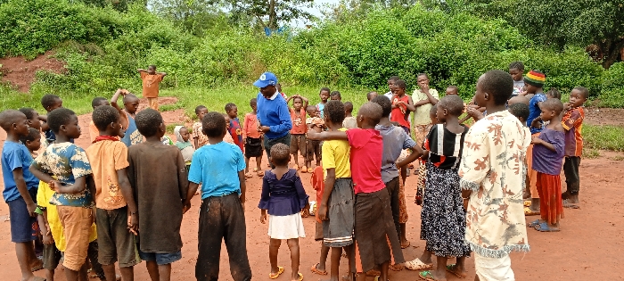 An outdoor scene in Ngalula, Democratic Republic of the Congo, depicts an adult male interacting with a group of approximately 25 to 30 children. The adult, positioned near the center of the frame, wears a blue long-sleeved shirt, dark pants, and a blue baseball cap with a white logo. He faces the children, with his right hand holding a small, light-colored object. The children, predominantly young, are gathered in a loose semicircle around the adult and extending towards the right. Their attire consists of various colored t-shirts, shirts, shorts, pants, and skirts, some appearing worn or soiled with reddish-brown dirt. Several children are barefoot or wear open-toed sandals. One child stands further back on a slight rise in the terrain, facing the main group with hands on hips. The ground is unpaved, reddish-brown dirt. The background is characterized by dense green foliage and trees, indicative of a natural or undeveloped environment. The interaction appears to be instructional or communicative, with children observing the adult.