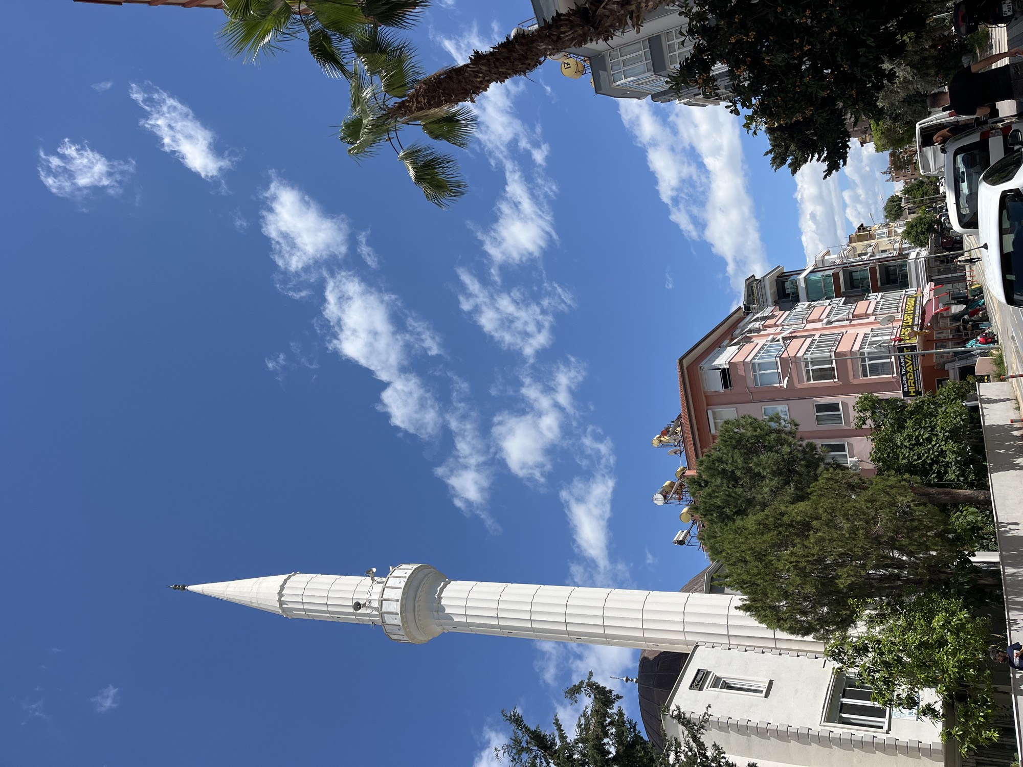 The image shows a low-angle view of a city scene under a blue sky with scattered white clouds. A tall, white minaret, likely part of a mosque, dominates the lower left. To the right, a mix of buildings can be seen, including a multi-story pink building and others with various architectural styles. Trees are interspersed among the buildings, adding greenery. Further to the right, a row of parked vehicles indicates a street. At the top of the image, a palm tree is visible, suggesting a warm climate. Based on the architectural style of the minaret and other buildings, as well as the presence of palm trees, the location is consistent with the city of Alanya, Turkey. There are no visible people or explicit actions or interactions occurring in the scene.