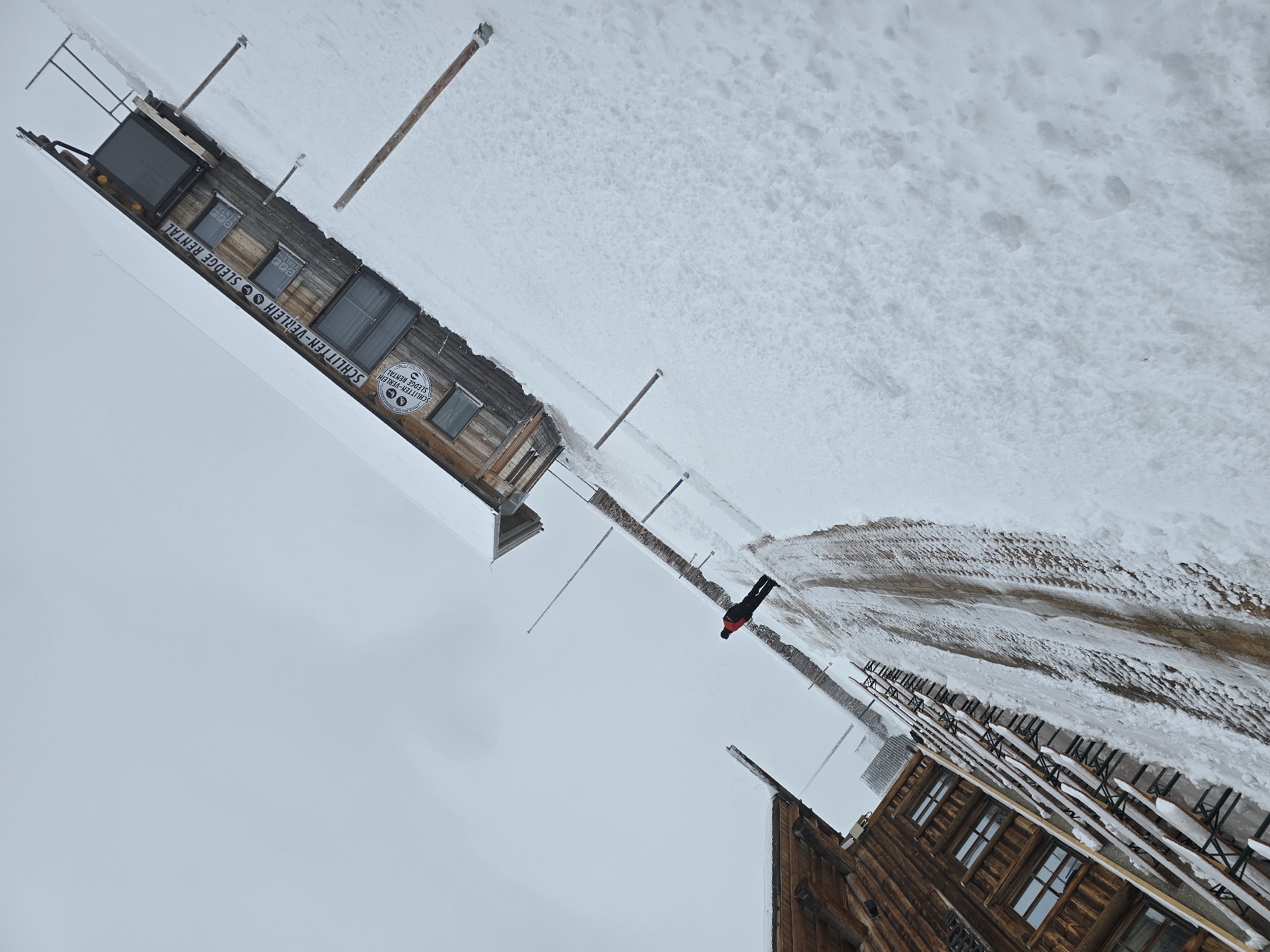 This media file captures a snowy winter scene in Garmisch-Partenkirchen, Germany, featuring wooden buildings and a lone person. The image is taken from a slightly elevated perspective, looking across a snow-covered landscape under an overcast sky.
In the upper portion of the frame, a rustic wooden building stands prominently, identified by a large sign across its facade that reads "SCHLITTEN-VERLEIH • SLEDGE RENTAL" in both German and English. A smaller circular sign on the building also displays "BOBS CLUB" with a sledder icon. Several wooden posts extend vertically from the deep snow surrounding this structure.
In the mid-ground, a single individual wearing a bright red jacket and dark trousers is walking on a path. The path, which appears to be a mix of snow and dirt, shows signs of being trodden or cleared.
In the foreground, another traditional wooden building with multiple windows is visible. In front of this building, numerous rows of wooden picnic tables and benches are arranged, each heavily blanketed with snow, suggesting they are out of use due to the winter conditions.
The scene conveys a calm, cold winter day in a mountainous region, characteristic of an alpine resort or winter recreation area, with clear indications of activities like sledding or bobsledding. The sky is a uniform gray, indicating an overcast day, and the ground is extensively covered in snow.