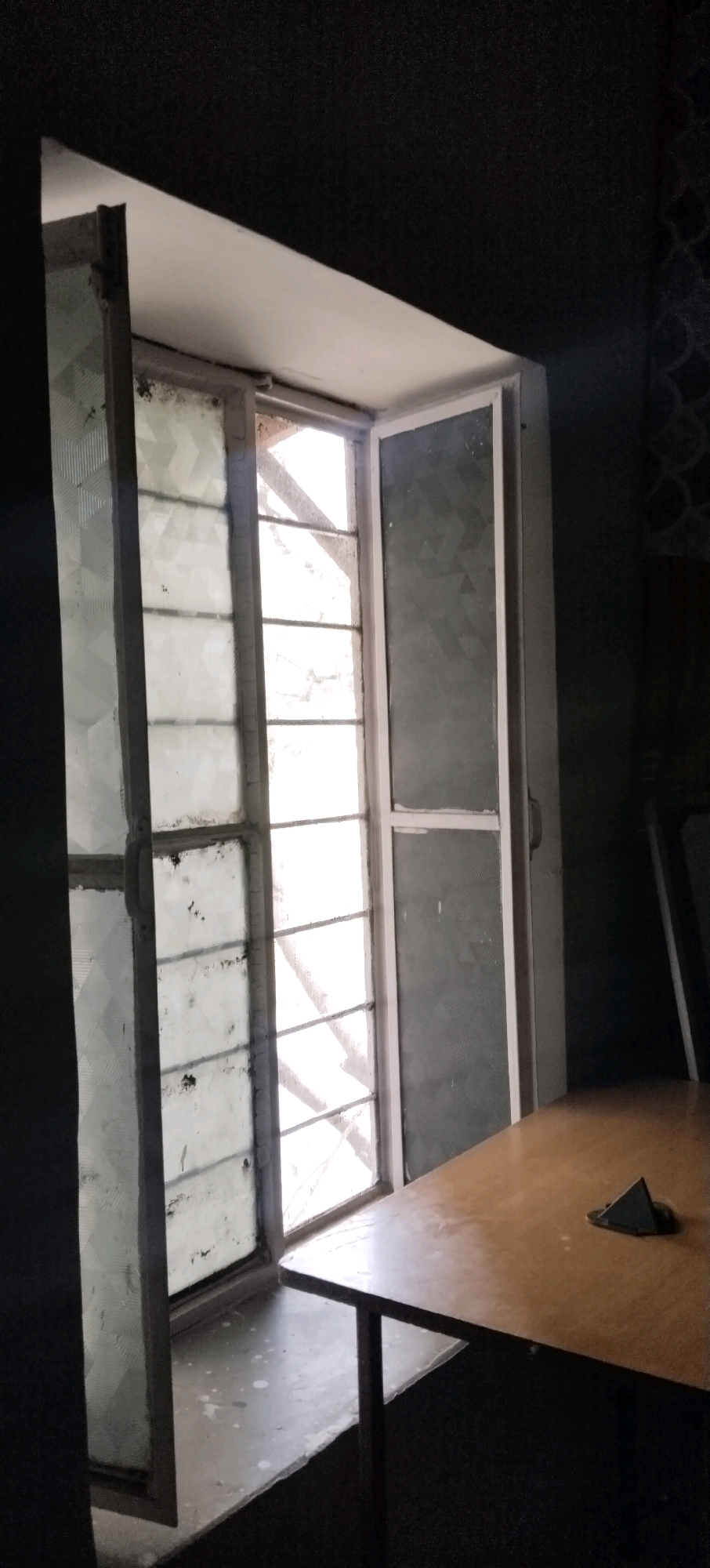 The image depicts a dimly lit room with a large window dominating the scene. The window is divided into multiple panes, with the left side of the window open, revealing a textured glass with a geometric pattern. The right side of the window is closed and also features textured glass. Sunlight streams through the window, illuminating the interior and casting shadows. A portion of a wooden table is visible in the foreground, with a small, dark, triangular object resting on its surface. The walls of the room appear dark, with one wall showing a subtle, possibly patterned texture. The overall impression is one of a functional, perhaps slightly dated, interior space. The location is specified as Mahapura, India.