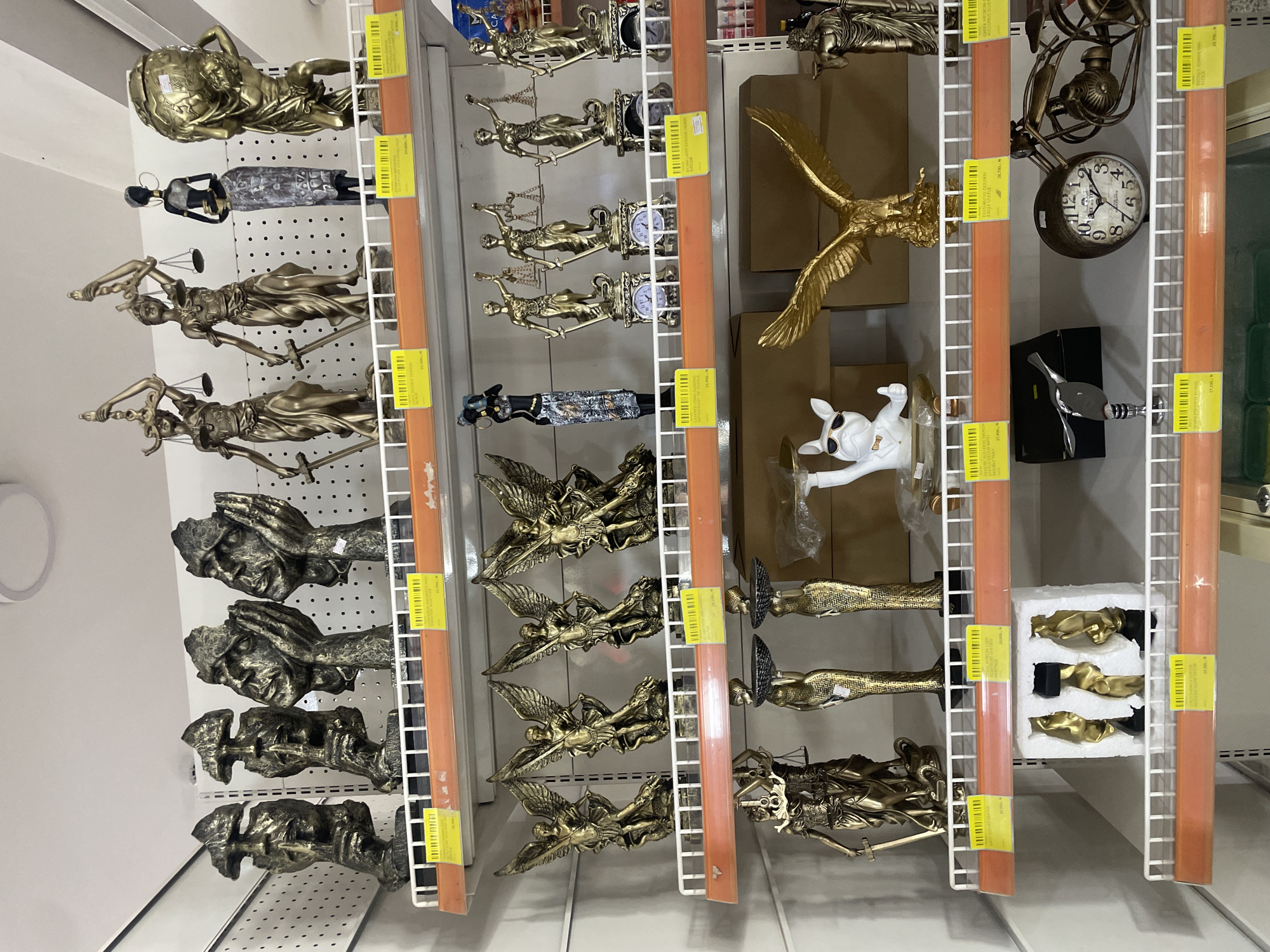 This is a medium shot of a retail display of various decorative statues and figurines, likely in a shop. The items are arranged on white wire shelves, with orange vertical supports visible. The overall color palette is dominated by gold and bronze tones from the statues, interspersed with the white of the shelves and the orange of the supports.
The subjects are primarily inanimate objects. On the left, there are several golden statues of female figures holding scales and swords, commonly representing Lady Justice. Below these are more abstract, dark-colored figures with hands covering their faces. Further down, there are more golden, winged figures.
In the center, a large golden eagle statue is prominently displayed. To its right, a white dog figurine wearing sunglasses and a bowtie stands out. Further to the right, a vintage-looking clock with Roman numerals is visible. Other items include metallic ducks and abstract golden forms.
The scene appears to be an indoor retail environment, possibly a home decor or gift shop. There are price tags visible on some of the items, with text that includes "GOLDEN," "EAGLE STATUE," "BULLDOG TR," and numerical values like "17.300 N," "28,700 N," and "900N." The location context provided states "Galadimawa Village, Nigeria," suggesting this is a shop in that region. There is no discernible activity or event taking place, and the image is static. The lighting is neutral and appears to be from overhead fixtures, indicating it could be daytime or evening.
Notable details include the metallic finishes of the statues, the variety of figures from classical representations to more modern and whimsical designs, and the presence of price tags indicating commercial activity.