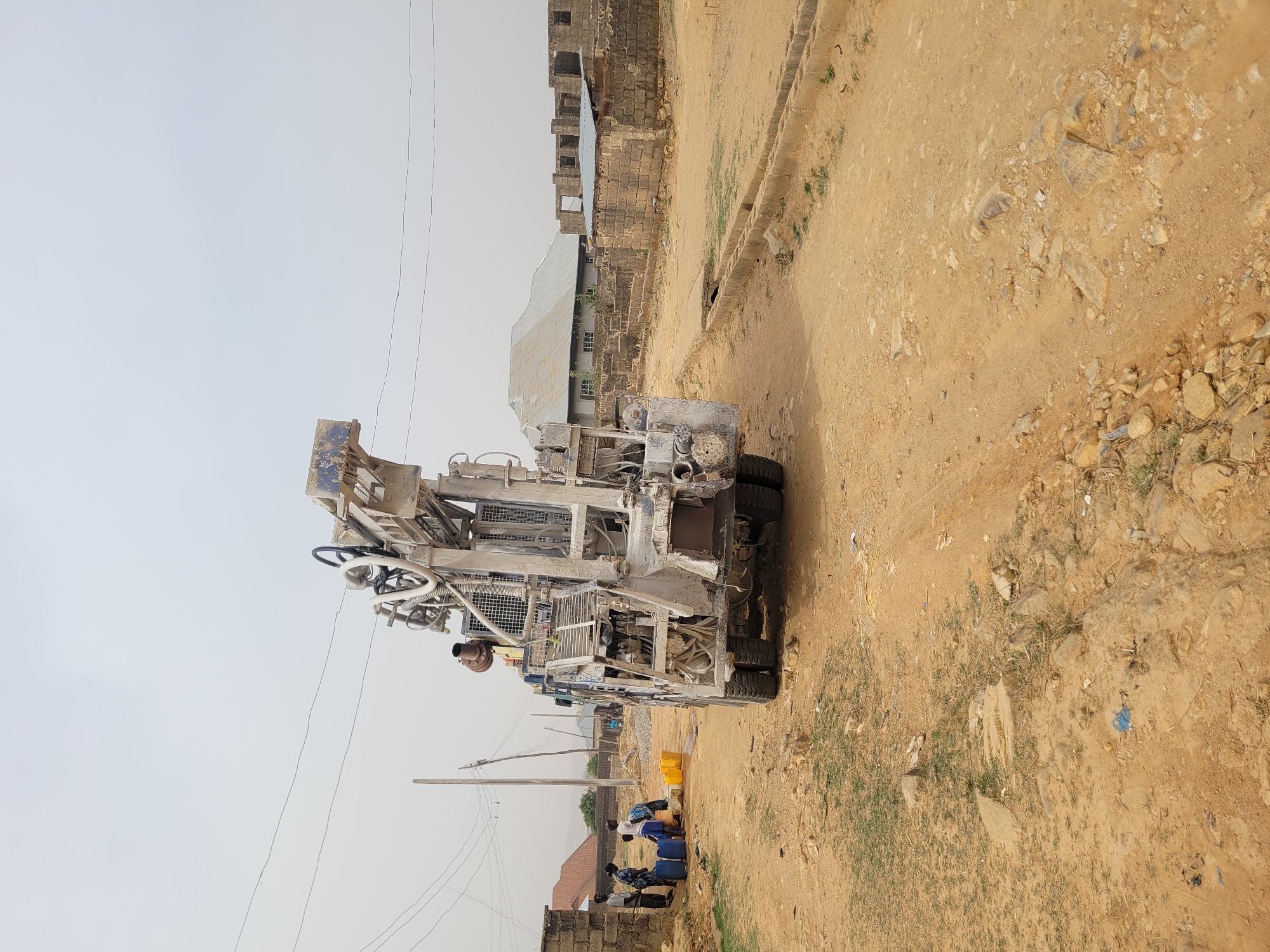 A truck-mounted drilling rig, painted in faded blue and grey and heavily coated in dust, is positioned on a dry, unpaved road in what appears to be a semi-urban or rural setting. The large piece of machinery dominates the left side of the frame, featuring a tall mast, various pipes, hoses, and mechanical components.
Several individuals, mostly men, are gathered near the rear of the drilling rig, on the ground to the right. They are seen around a collection of blue and yellow plastic jerrycans or drums, suggesting activities related to water collection or handling, possibly in conjunction with the drilling operation or for local use. One person in a blue shirt is particularly visible amongst the group.
The surrounding environment is characterized by dry, sandy-brown earth with scattered small rocks and sparse patches of green grass. To the right of the rig, an unpaved path runs alongside a concrete drainage channel. In the background, a cluster of buildings can be seen under a hazy, light grey sky. Some structures appear to be residential, featuring corrugated metal roofs, while others are incomplete, showing exposed block walls. Utility poles with overhead wires are also visible in the distance. The overall impression is of a construction or resource development site in a dusty, developing area. No legible text is visible on the rig or in the surroundings. The time of day appears to be daytime under an overcast or hazy sky.