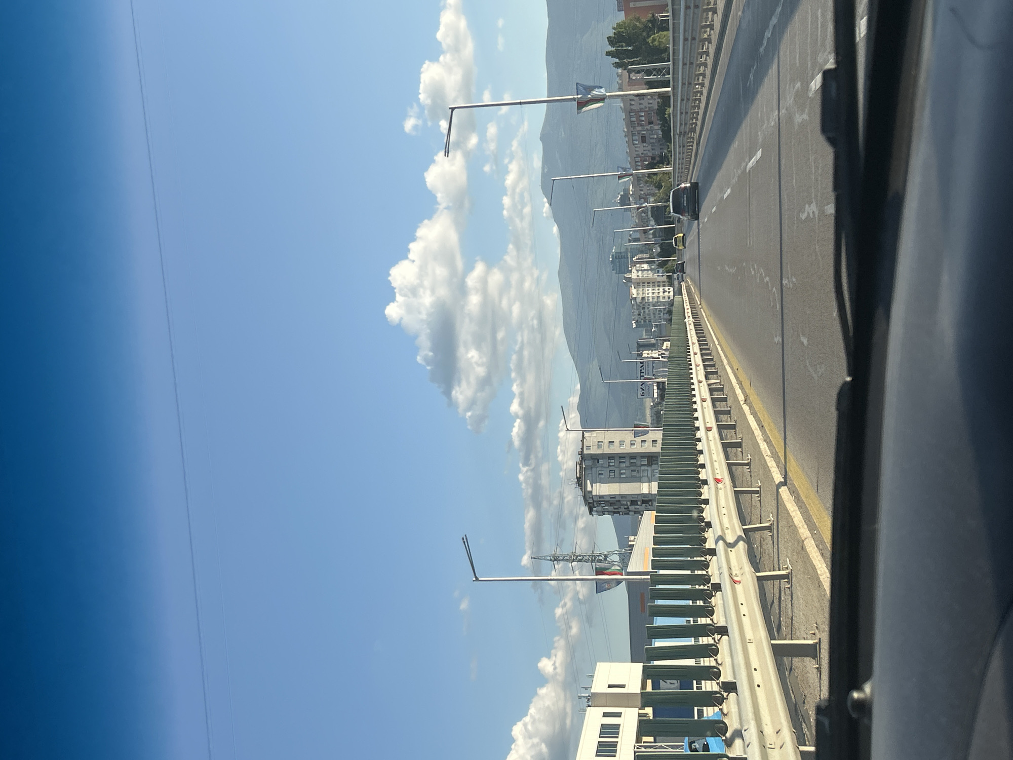 The image depicts a view from a vehicle traveling on a highway overpass in Sofia, Bulgaria. The overpass has a guardrail on the right and a metal fence with green panels on the left. Multiple streetlights line the highway. In the distance, apartment buildings and industrial structures are visible, with a large mountain range serving as a backdrop. The sky is clear blue with scattered white clouds, and the sun appears to be setting, casting a warm glow. Several cars are driving on the highway.