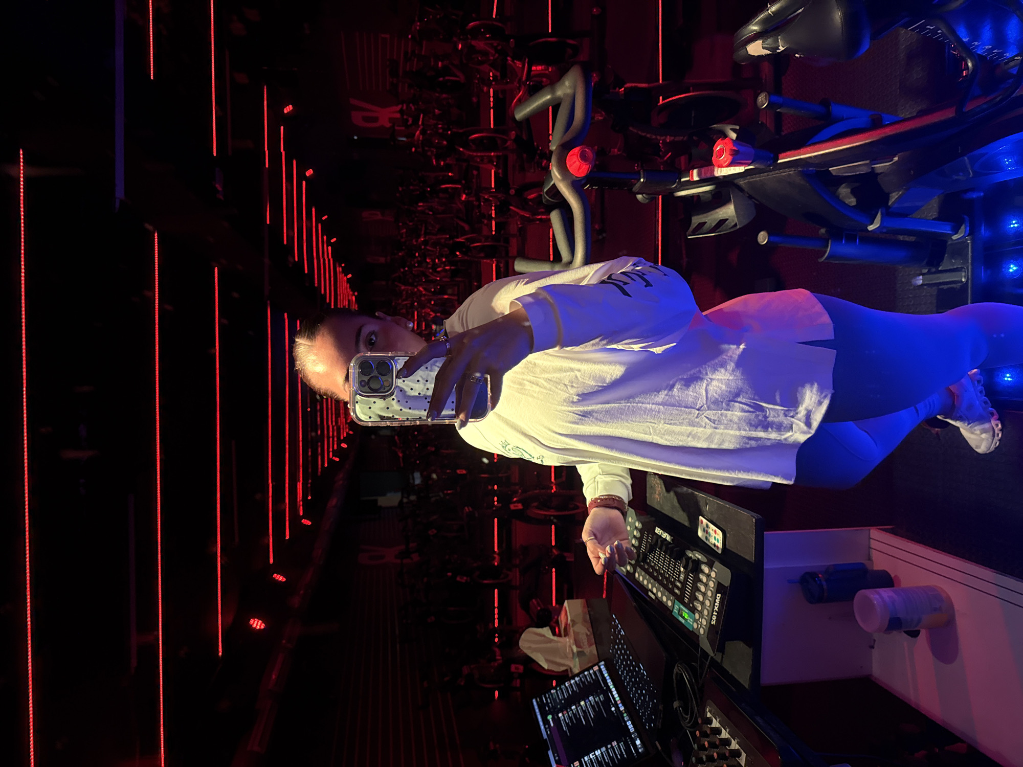 A woman takes a selfie in a cycling studio featuring exercise bikes, red vertical lights, and control equipment. She holds a smartphone in her right hand, wearing a white long-sleeve shirt and blue leggings. Exercise bikes are arranged in rows behind her, illuminated by the red lights. A control panel and a laptop computer are visible on a surface adjacent to the woman. The studio also includes blue lights on the right. There are no indications of a precise location in Puebla, Mexico.