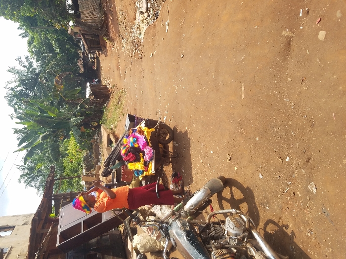 The scene is an unpaved reddish-brown dirt road in the city of Nnobi, Nigeria, captured during daylight. In the left foreground, a dark, weathered motorcycle is parked on the dirt. Adjacent to it, an adult female stands barefoot, wearing an orange short-sleeved top, maroon trousers, and a brightly colored, multi-patterned conical hat. She holds a small rectangular mirror in her hands, looking into it. Beside her is a two-wheeled handcart loaded with numerous brightly colored fabric items, predominantly yellow, pink, purple, and blue. Behind the woman and cart, an incomplete concrete block building rises, with dense tropical foliage, including large banana plants, visible beyond. The dirt road extends into the background, flanked by various simple structures with corrugated metal roofs and additional green vegetation, before curving slightly to the right and receding into the distance. Scattered debris is present on the road surface.