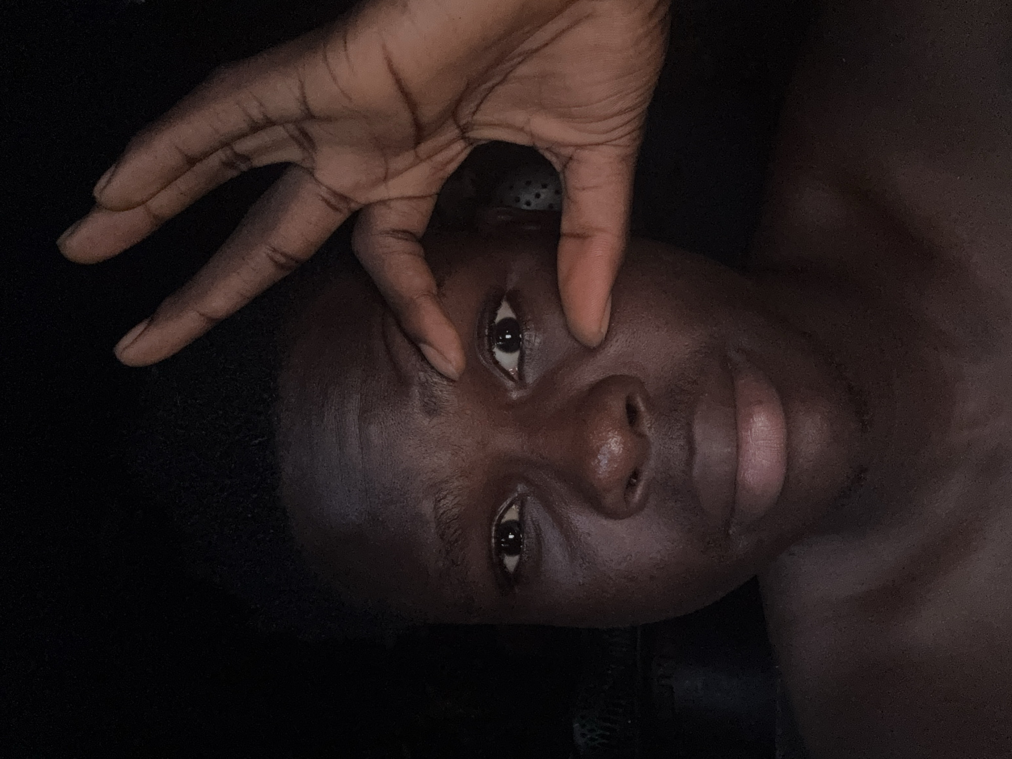 A dark-skinned person lies on a dark surface. A dark-skinned hand with fingers extended is above the person's face, with the index finger touching the upper eyelid of the person's right eye. The person's face is facing the camera, and their eyes are open. The background is dark and undefined. The image provides no information indicating a location in Oboama, Nigeria.