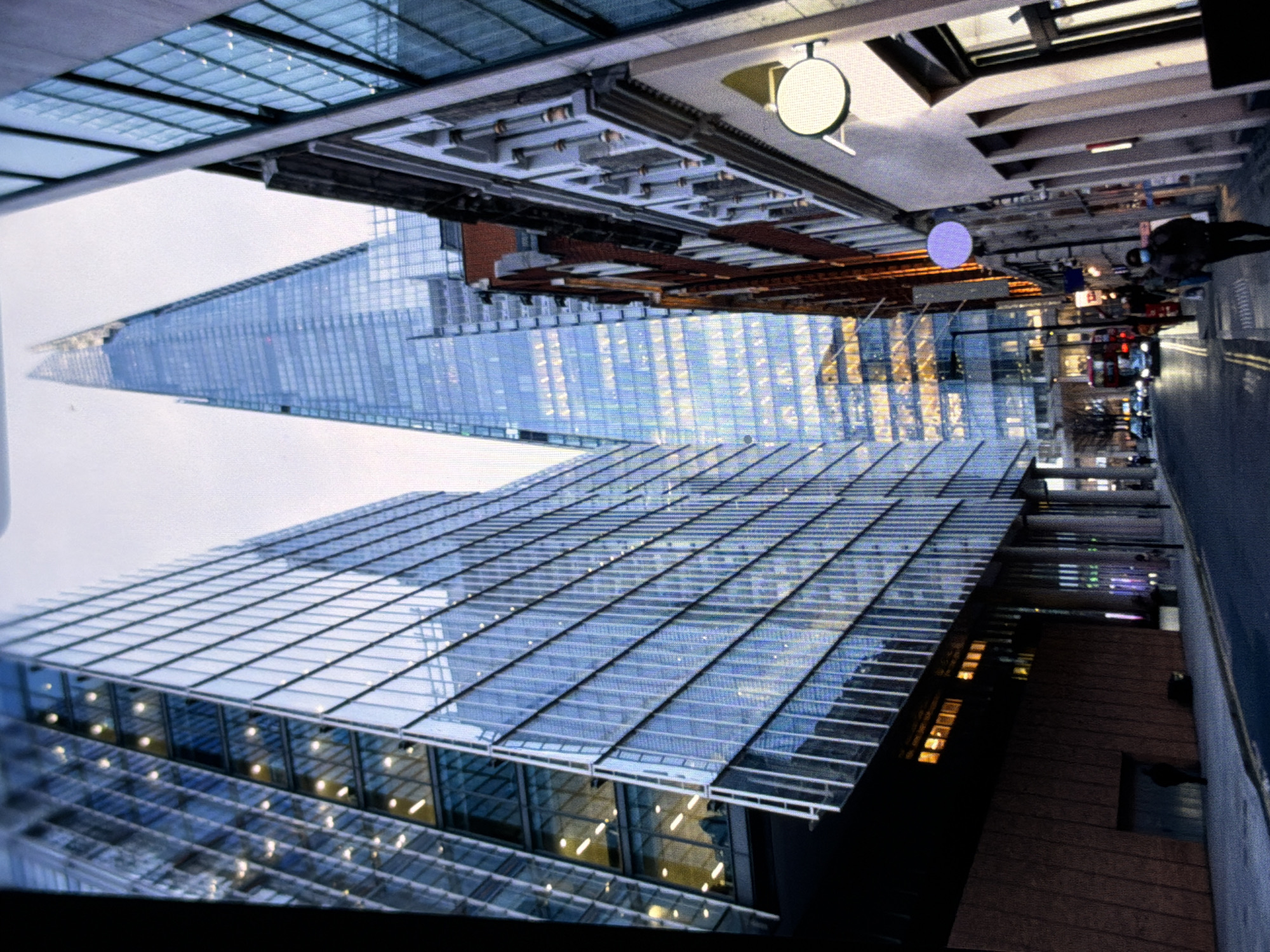 This image depicts a street scene in London, United Kingdom, not Maidstone, prominently featuring The Shard skyscraper.
In the background, The Shard, a tall, glass-clad pyramidal skyscraper, stands centrally against an overcast, light grey sky. On the left, a multi-story modern building with a blue glass curtain wall displays numerous illuminated interior office spaces. Its lower section features a dark, textured facade. On the right, a sequence of buildings includes a structure with a light-colored, decoratively carved upper facade and a plain lower section, from which a circular white sign projects. Further along this street, a building with a red brick facade is visible, above which a circular purple light is mounted.
The foreground shows an asphalt street with visible double yellow lines, flanked by concrete pavements. On the right-side pavement, a pedestrian wearing a dark coat and a blue face mask walks towards the viewer. Additional pedestrians are discernible further down the street. Vehicular traffic, including a red double-decker bus and several cars with illuminated headlights, is present in the middle ground. Multiple streetlights are active. The ambient lighting from the sky is subdued, consistent with an overcast day or early evening.