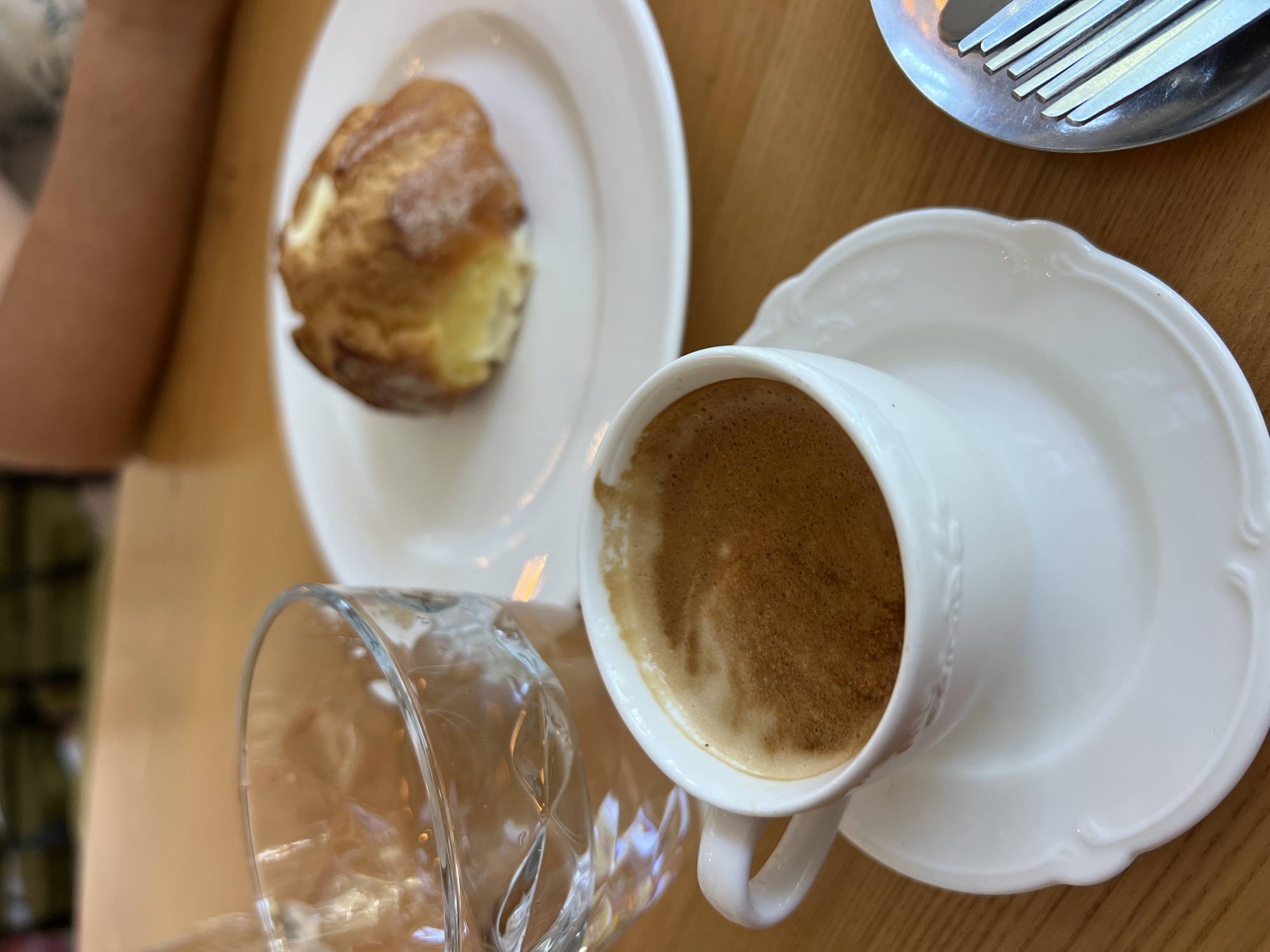 A close-up, eye-level shot shows a white espresso cup and saucer on a wooden table. The espresso has a thick layer of crema. To the left of the cup is a glass of water with ice. In the background, on a white plate, is a pastry, possibly a brioche or a choux pastry. A person's arm is visible in the upper right corner of the frame, and part of a fork is seen on a small plate in the lower right. The overall impression is that of a casual cafe setting, possibly in Odesa, Ukraine, given the context.