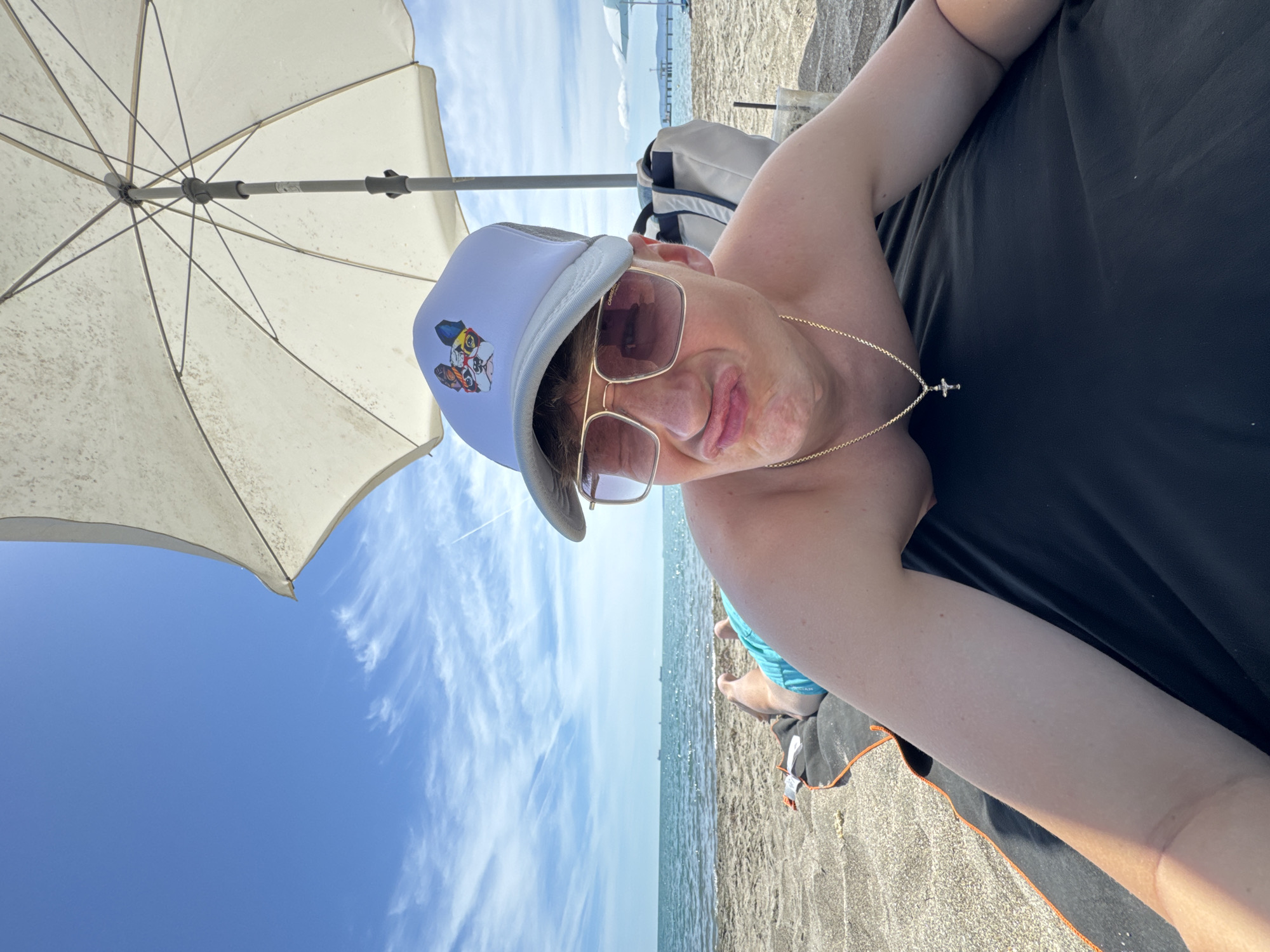 A young person is lying on a beach towel on the sand, under a white umbrella. The person is wearing a white trucker hat with a dog graphic, sunglasses, and blue swim trunks. They are making a funny face, puckering their lips. The ocean and sky are visible in the background. The location is the beach in Burgas, Bulgaria.