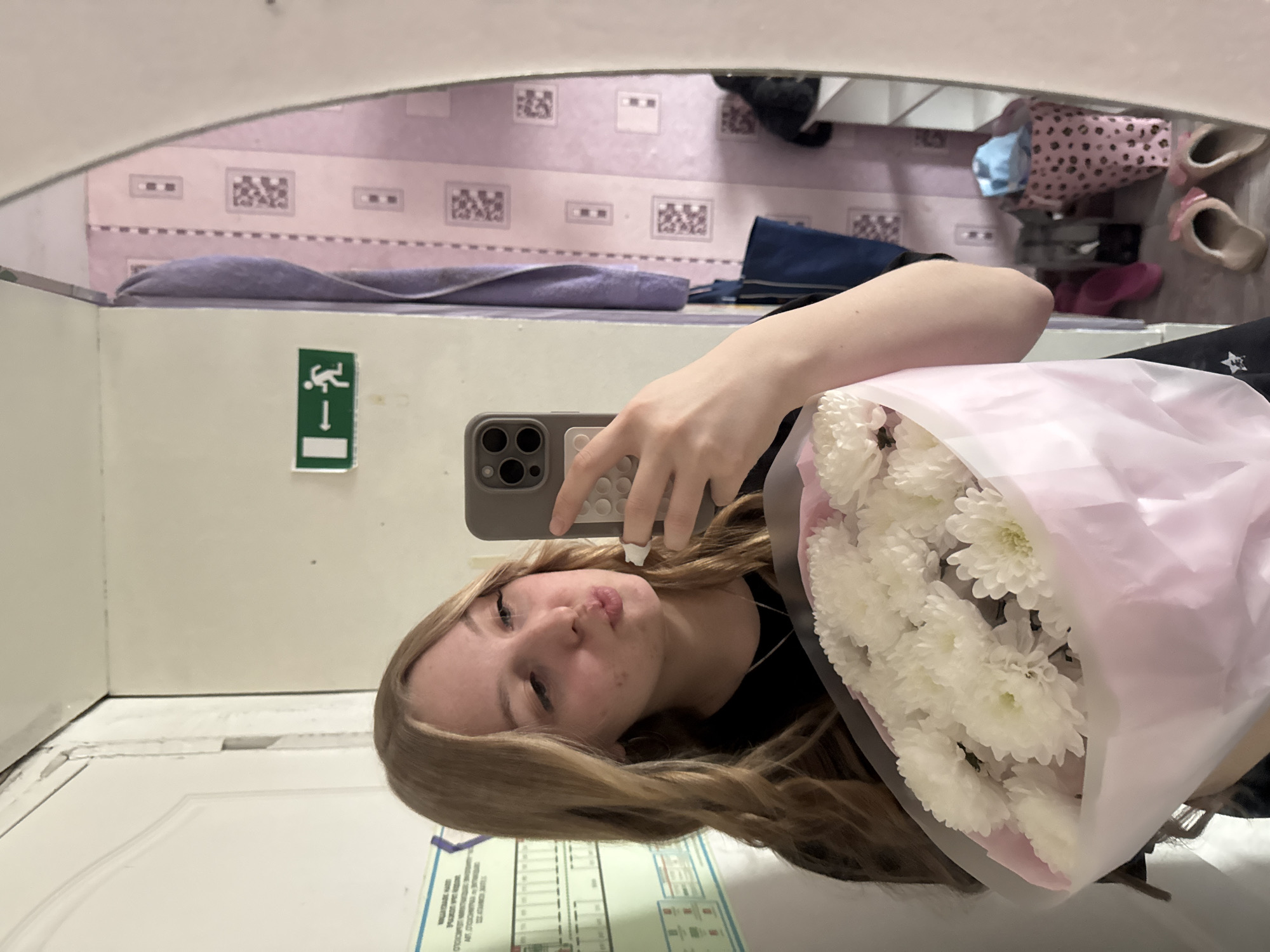 The image is a mirror selfie taken by a young woman, presented rotated 90 degrees clockwise. The woman, with long, wavy blonde hair, is shown from the chest up, making a playful pouting expression as she looks into the phone held in her right hand. She wears a dark top. In her left arm, she holds a large, fresh bouquet of white chrysanthemums, wrapped in clear and delicate pink cellophane.
The background, visible as a reflection in the mirror, shows an indoor setting, likely a room or hallway. A wall with pinkish-purple patterned wallpaper featuring lighter square designs is prominent. Below this, a purple towel is draped over a shelf or ledge. To the right, various personal items are scattered, including a pink leopard-print bag, a pair of light-colored slippers adorned with pink bows, and another pair of pink shoes or sandals.
On the wall closer to the foreground, a green emergency exit sign with a white pictogram of a running figure and a downward arrow is clearly visible. Further down and to the left, a white door or wall section is present. In the bottom-left corner of the reflection, a white document or poster with multiple lines of text and diagrams is affixed, though the text itself is too small to be legible.
The overall scene suggests a casual, indoor environment, possibly a home or private space in Perm, Russia. The woman's expression and the flowers imply a cheerful and lighthearted moment.