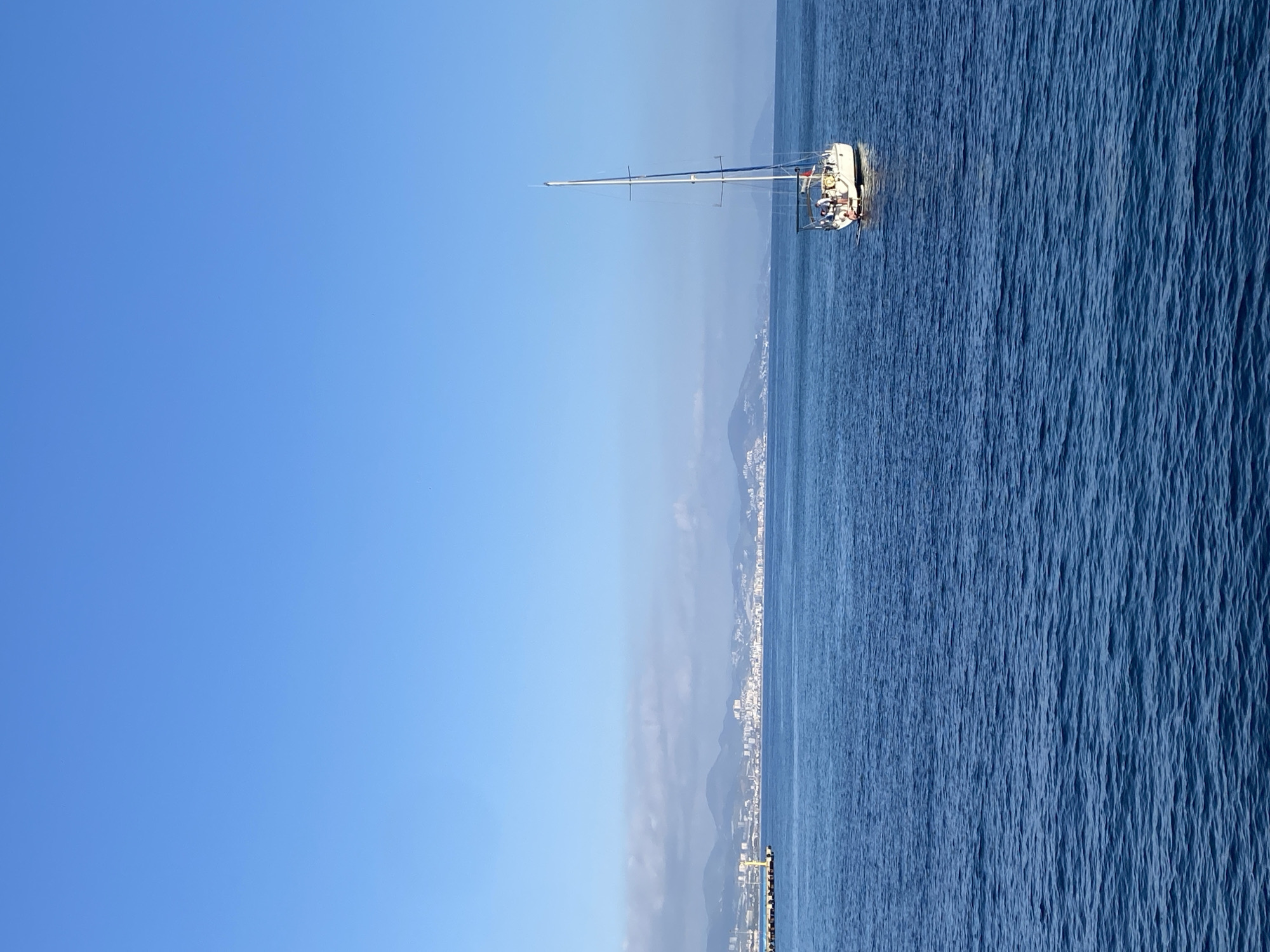 A serene daytime scene unfolds with a white sailboat gracefully positioned on calm, blue waters under a vast, clear sky. The sailboat, featuring a prominent tall mast and rigging, appears to be at rest or moving gently. Several people are discernible in the boat's cockpit, suggesting a leisurely excursion.
The expansive blue sea exhibits subtle ripples and texture across its surface, catching the bright sunlight and varying slightly in shade. Above, the sky is a brilliant, cloudless azure, indicating fair and sunny weather conditions.
Along the distant horizon, a long stretch of coastline is visible, featuring mountains that meet the sea. Urban development, characterized by numerous light-colored buildings, is evident along the base of these mountains, consistent with a coastal city like Alanya, Turkey. A slight atmospheric haze softens the details of the distant land. The overall mood conveyed is one of peace and tranquility on a beautiful day at sea. No visible text is present in the image.
