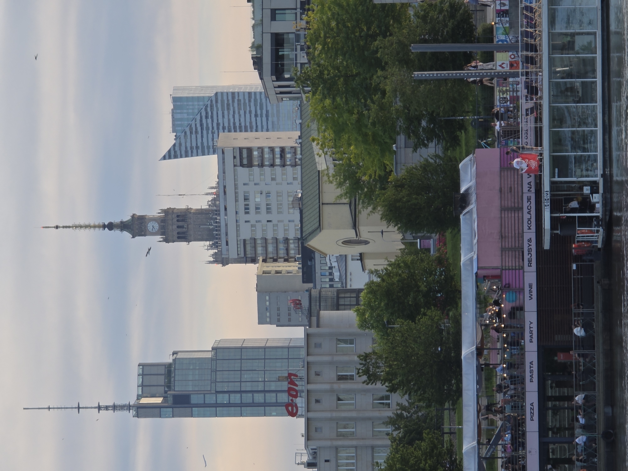 The image depicts a cityscape of Warsaw, Poland, dominated by modern and historical architecture. In the foreground, a floating restaurant or bar on the Vistula River is visible, with people dining and socializing on its upper deck. The facade of the floating structure displays text indicating "PIZZA," "PASTA," "PARTY," and "WINE." Behind the riverfront, a line of buildings forms the city skyline. Prominent structures include a tall skyscraper with an antenna on top, another distinctive skyscraper with a sharp, angled facade, and a building with a clock tower, likely the Palace of Culture and Science. The sky is overcast with a soft, diffused light, suggesting it is either early morning or late afternoon. Trees are interspersed between the buildings, adding a touch of greenery to the urban landscape. Several birds are visible in the sky, and a small airplane can be seen in the distance.