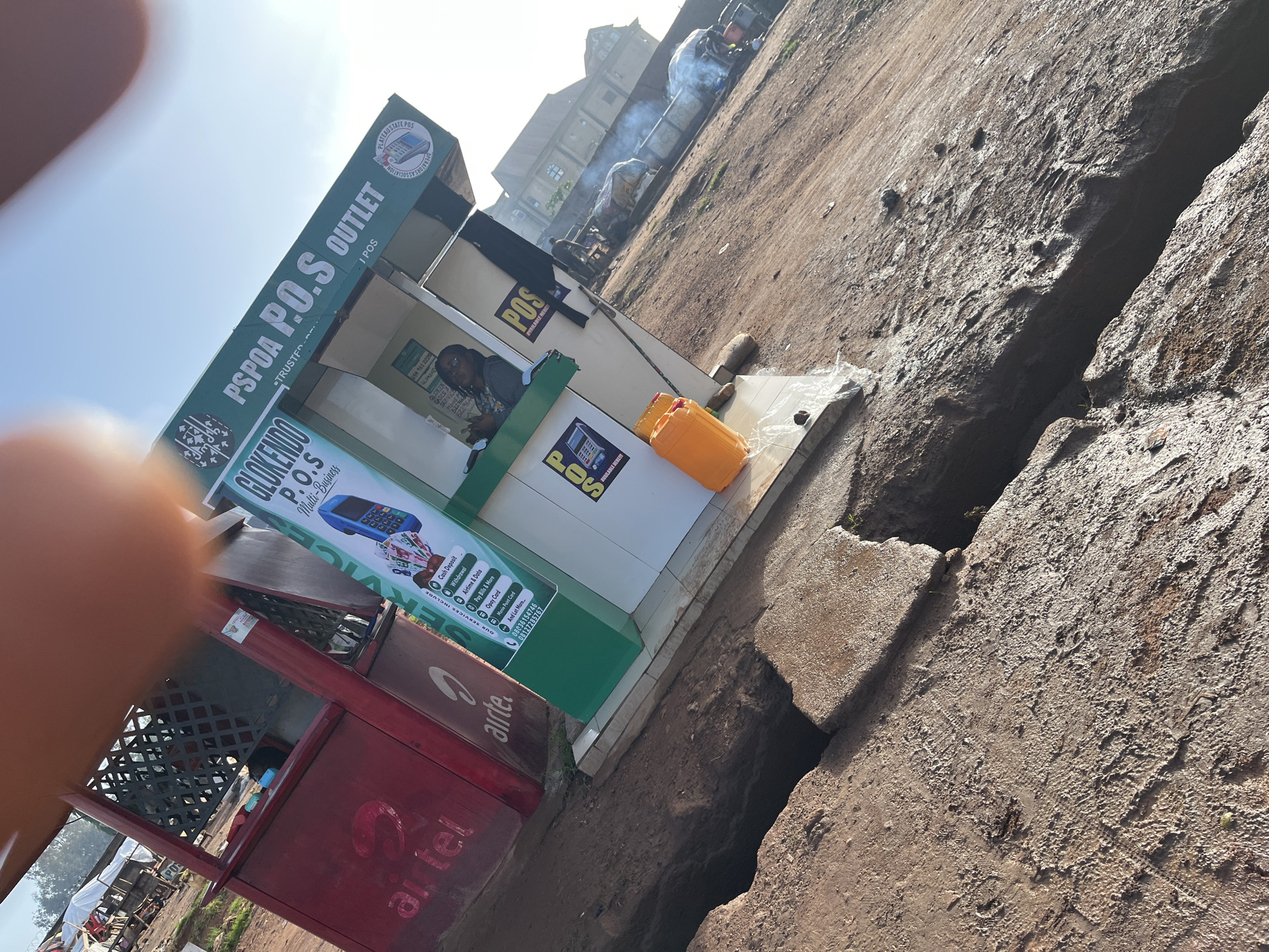 The image captures a bustling P.O.S. (Point of Sale) outlet in Gura Top, Nigeria, on a bright, clear day. The outlet is identifiable by a green and white booth displaying "PSPOA P.O.S. OUTLET" and "GLOKENDO P.O.S. Multi-Business" signage. Several services are listed, including "Cash Deposit", "Withdrawal", "Airtime & Data", and "Pay Bills & More". An Airtel branded kiosk is situated to the left of the P.O.S. outlet. A woman, likely an attendant, is visible inside the booth, handling transactions. Two orange containers are placed outside the booth. In the background, a building with arched windows and tiled roofs is visible, and further back, smoke rises from what appears to be a vehicle or machinery, suggesting industrial or commercial activity. The ground is a mix of cracked earth and paved areas, with some debris scattered around, indicative of an outdoor urban environment.