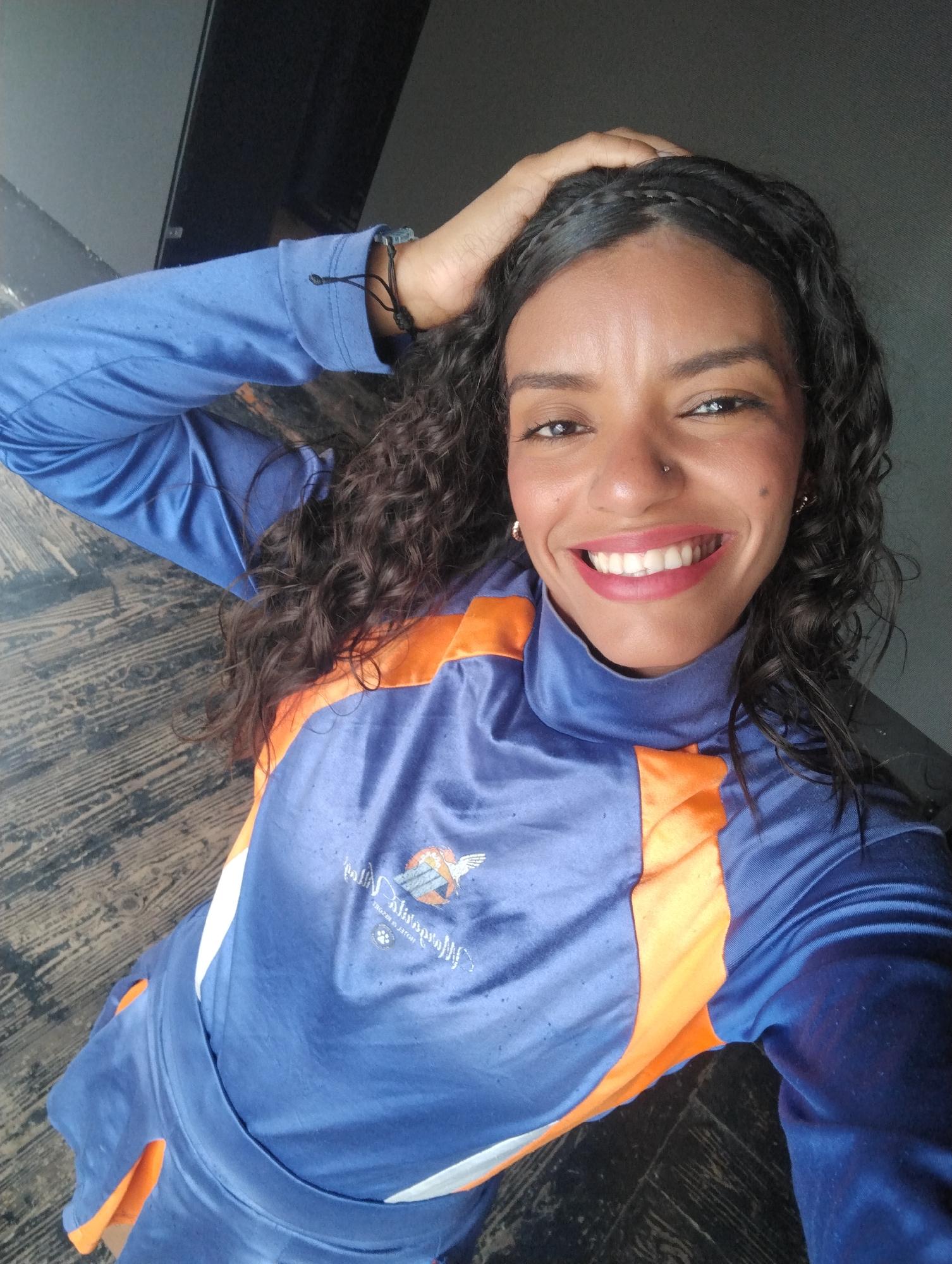 A dark-haired woman smiles in this overhead selfie. She has dark curly hair and wears a blue and orange tracksuit, her left hand on her head. She has dark eyebrows, a nose piercing, and wears red lipstick. The background is a dark grey wall and a brown wood-patterned surface. This is likely an indoor environment. There's no information in the image that points to Porlamar, Venezuela.