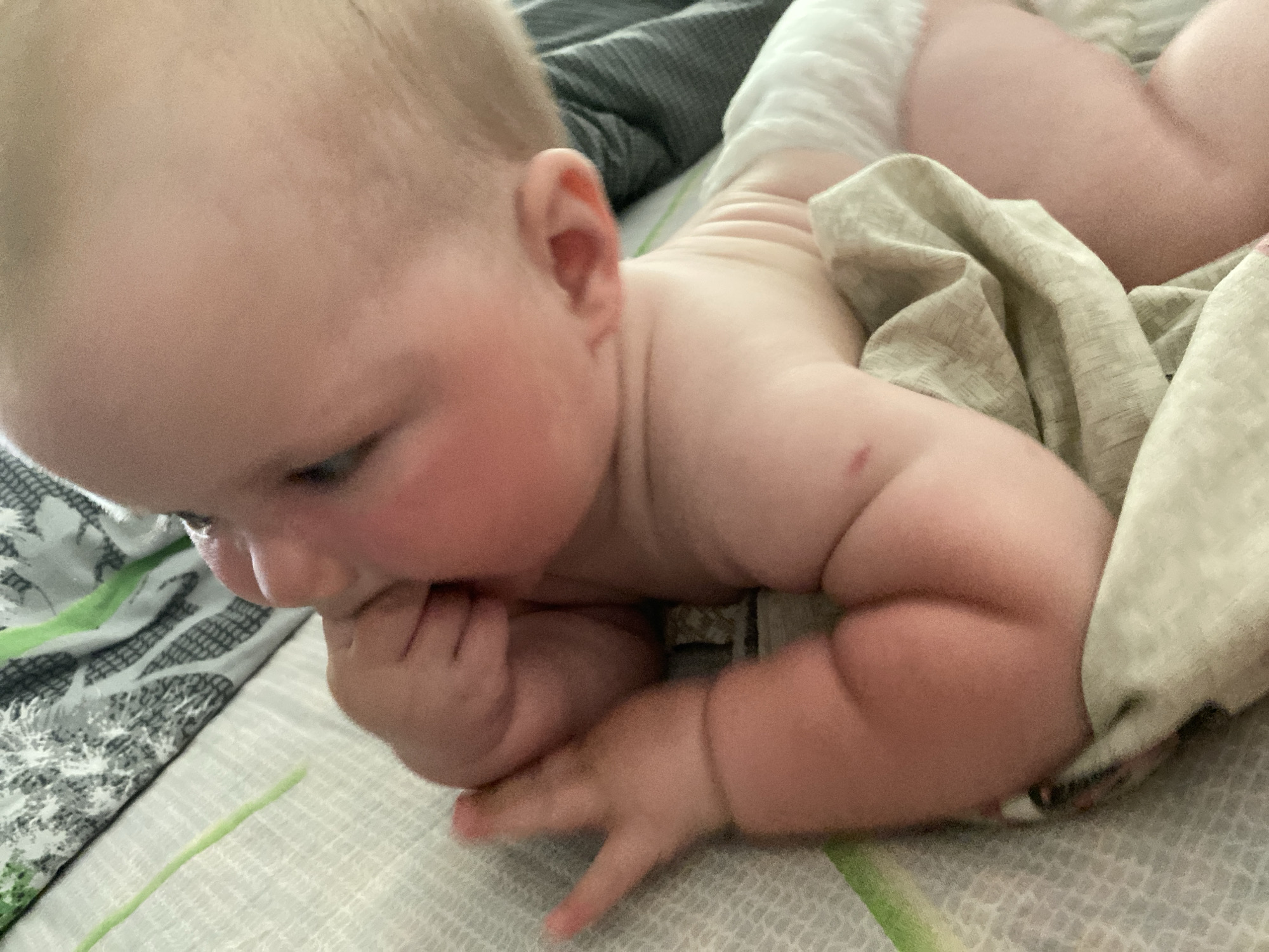 A close-up, top-down, slightly angled shot shows a baby lying on its side on a bed. The baby's head is turned away from the camera, with its face obscured by its hand, which is in its mouth. The baby has rosy cheeks and blonde hair. It is wearing a diaper. The bedding appears to be a light-colored patterned sheet and a darker grey blanket. A light green and white striped object, possibly a blanket or part of the bedding, is visible in the upper left corner and extends down the left side of the image.