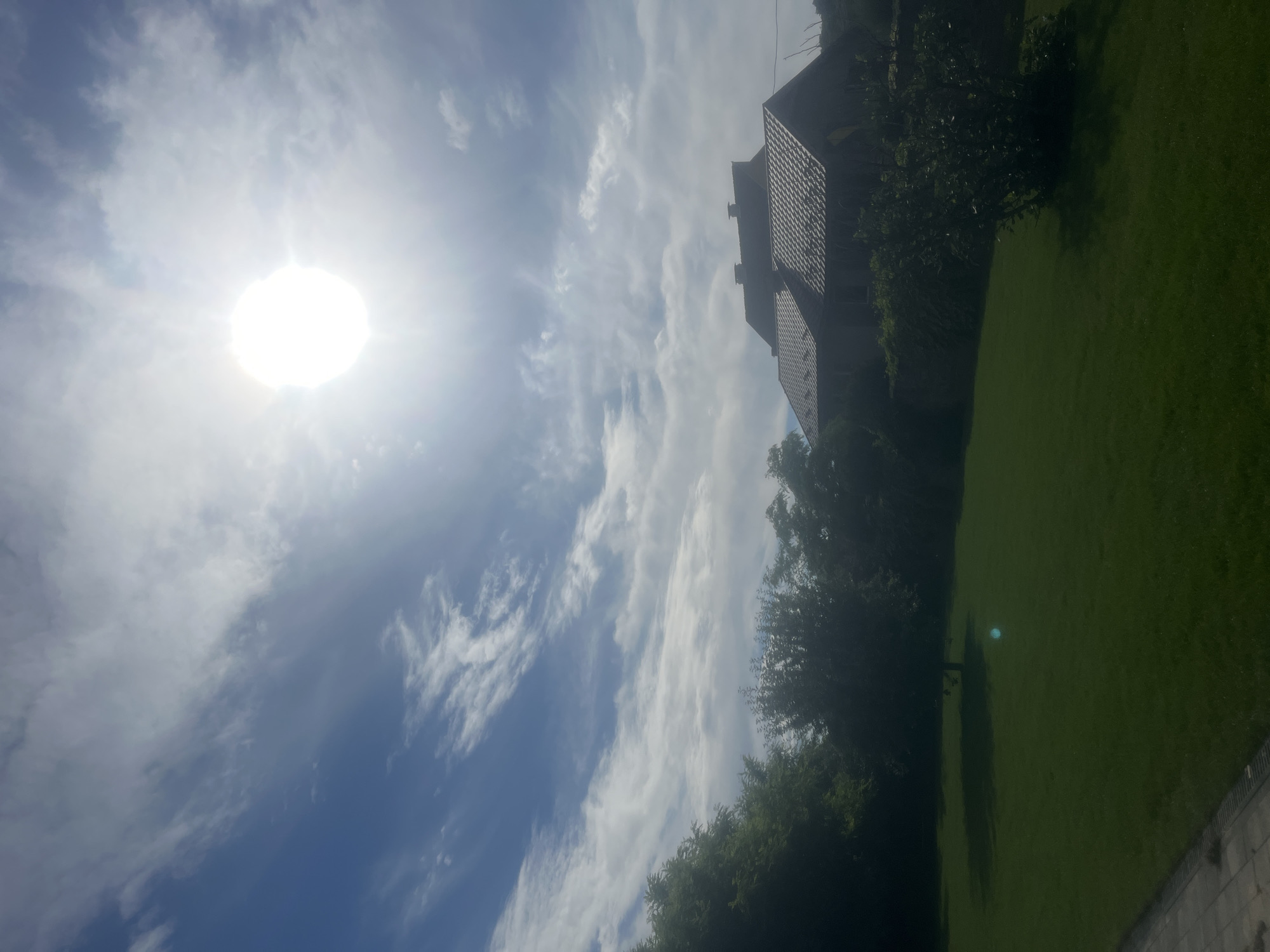 The image shows a bright, sunny day with a clear blue sky dotted with wispy white clouds. The sun is prominently visible in the upper right quadrant of the frame, emitting a strong glare. In the mid-ground, a two-story house with a tiled roof is visible, partially obscured by trees. The house appears to be situated in a suburban or rural setting, with a well-maintained lawn in the foreground. Trees and foliage surround the property, creating a green and natural environment. A paved pathway is visible along the bottom left edge of the image. There are no people or animals present in the scene. The overall impression is one of a peaceful, domestic setting bathed in natural light. This scene is located in the village of Onokivtsi, Ukraine.
