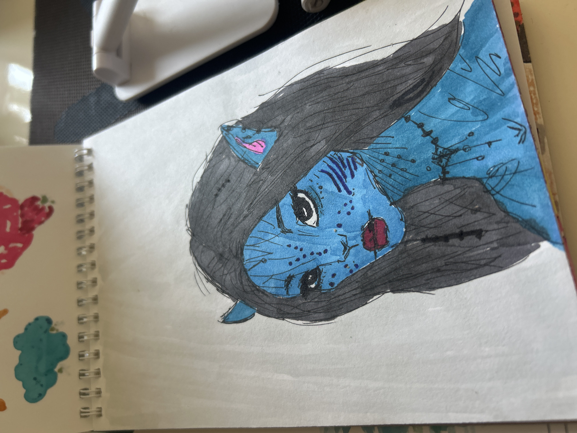 The image shows a drawing of a Na'vi character from the movie Avatar. The character has blue skin, black hair, and pointed ears with pink inner ears. They are depicted with tribal markings on their face and neck, and are wearing a necklace. The drawing is done in a sketchbook with a spiral binding. In the background, a white object, possibly a charging station or a piece of technology, is visible. The drawing appears to be on a table, with a hint of a patterned surface beneath the sketchbook. There is no indication of the city of Khyrdalan, Azerbaijan, in the image.