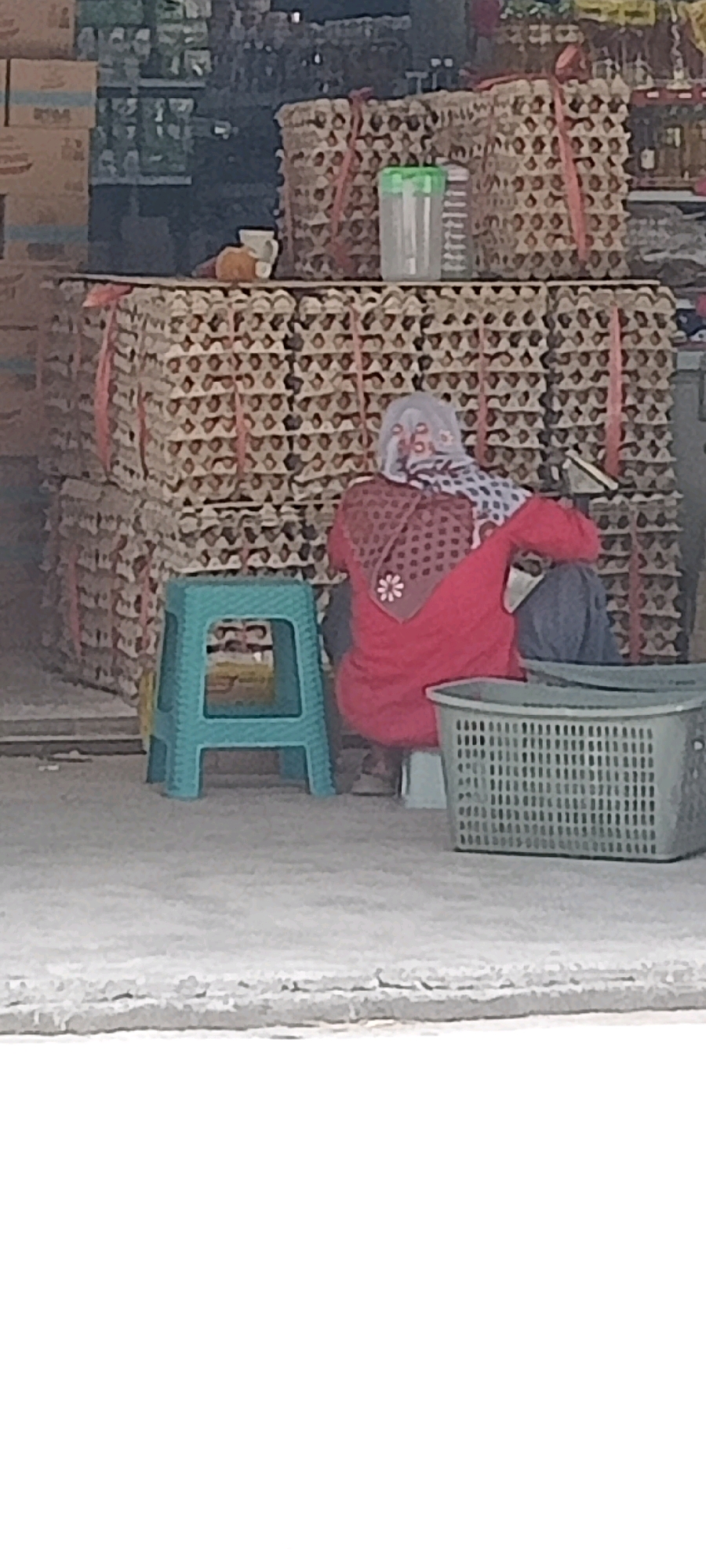 The image depicts an outdoor market stall in Desa Karangmojo, Indonesia, likely a vendor selling eggs given the stacked egg cartons. A person, seen from behind and wearing a headscarf and a red garment, is seated on a small white stool behind a blue plastic stool. They appear to be resting or waiting for customers. Several large stacks of brown egg cartons are visible, tied with pink ribbon. A grey plastic basket is placed to the right of the seated person, and in the background, there are shelves with various items, suggesting it's part of a larger shop or market. The ground is a rough, concrete-like surface.