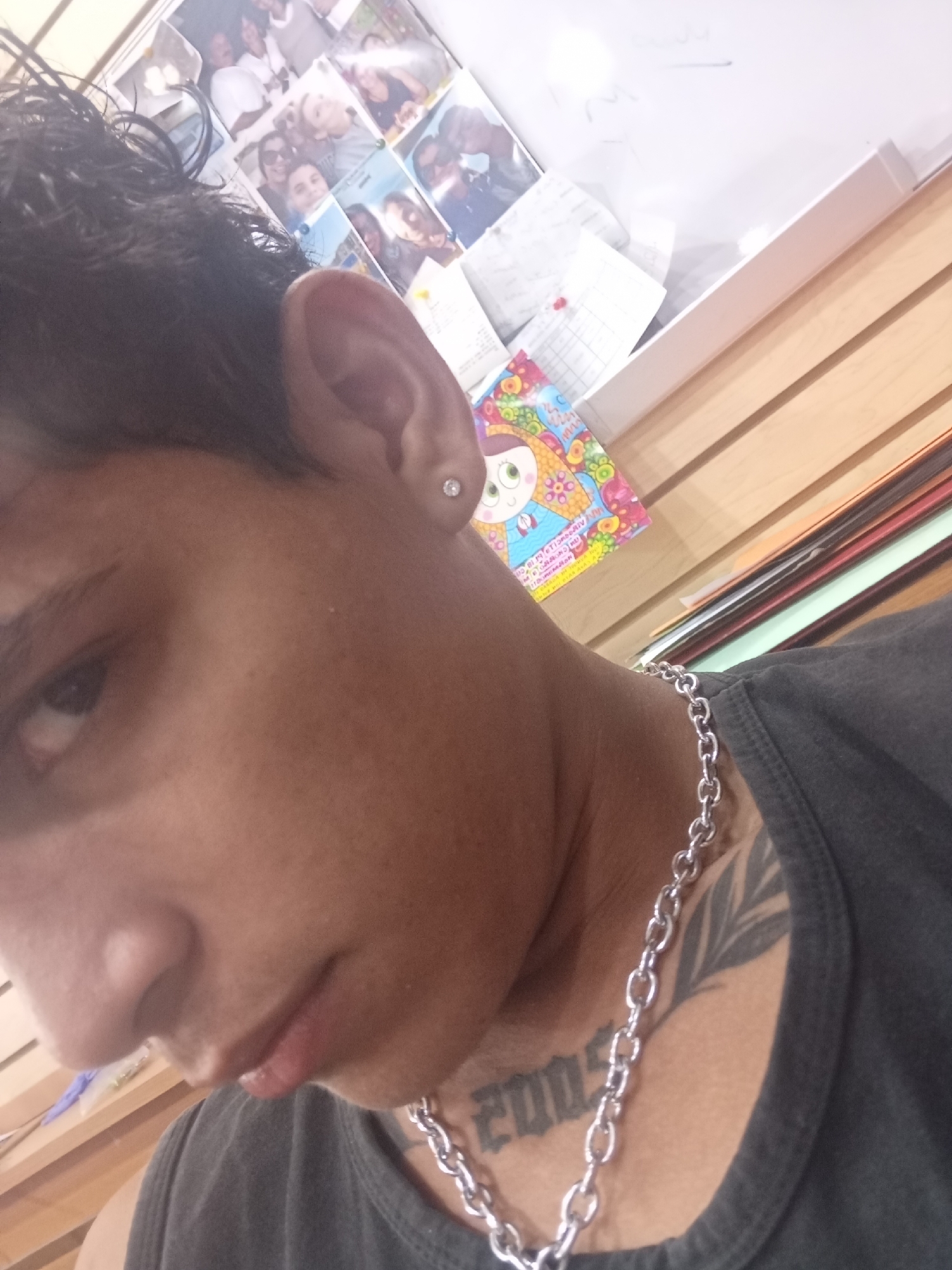 A close-up, high-angle photograph focuses on a person's head and upper torso. The person has short, dark hair and dark eyes. A stud earring is visible in the person's left ear. The person wears a silver chain necklace, and a tattoo of what seems to be a series of digits can be observed beneath the necklace. An additional tattoo of foliage is on the upper left chest, by the person's neck. The person wears a dark grey tank top.
In the background, a collage of photographs appears to be pinned to a whiteboard. A colorful, patterned object is visible in front of the whiteboard. Below these items, a stack of papers of various colors is seen on a surface with a wood-like pattern.
There is insufficient evidence to identify that this photo was taken in Juan Griego, Venezuela.