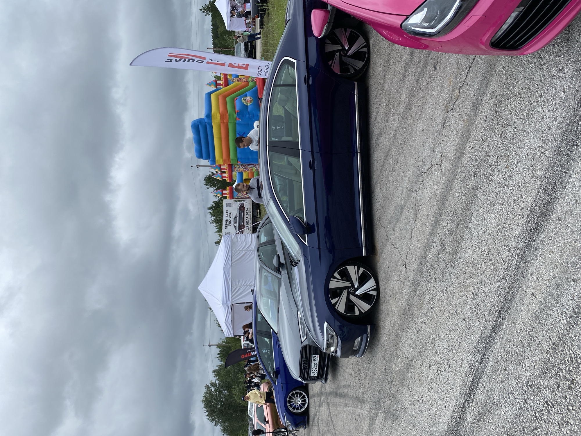A group of modified cars are parked in a line on a paved surface. The cars are of various colors, including dark blue, grey, and pink. Several people are standing around the cars, some of whom appear to be looking at the vehicles. In the background, there are white tents and a colorful inflatable playground. A flag with Russian text is visible on the right side of the image. The sky is overcast. The cars appear to be at an outdoor event, possibly a car show or festival. The license plate of the grey car reads "P382KK 159". This suggests the location could be within the city of Perm, Russia, as Russian license plates typically follow this format and the 159 region code corresponds to Perm Krai.