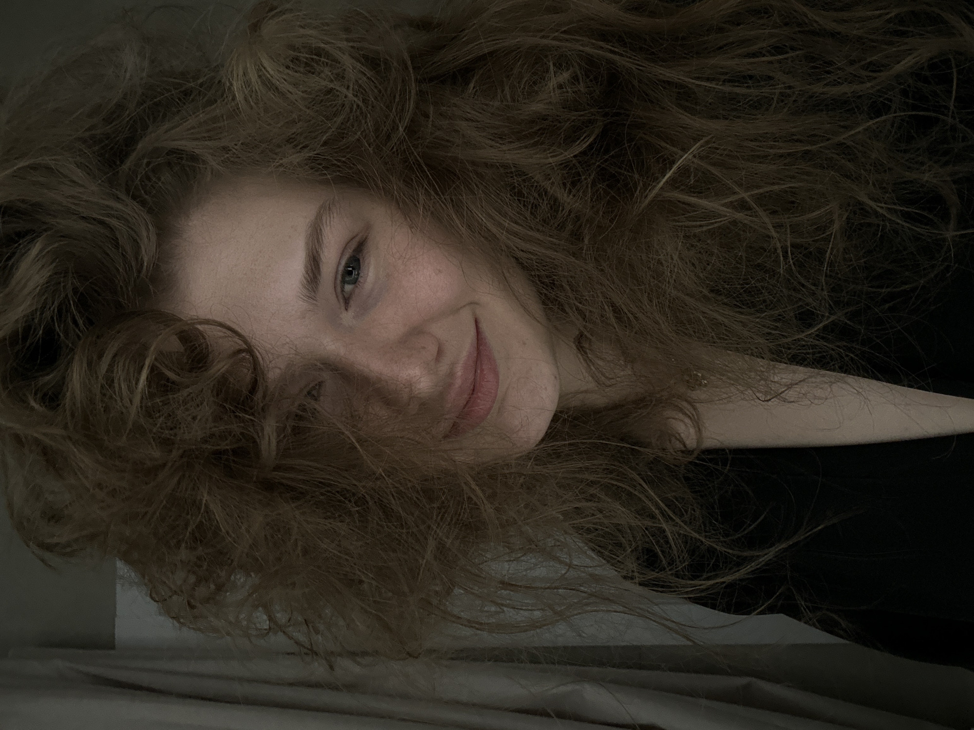 A young woman with light skin and auburn, wavy hair is lying on a bed, looking towards the camera with a slight smile. Her right side is visible, showing a portion of her neck and shoulder, covered by a dark-colored garment. The scene appears to be indoors, likely a bedroom, with a bed and light-colored sheets. The lighting is dim and diffuse. There is no clear evidence to suggest the location is Kazan, Russia.